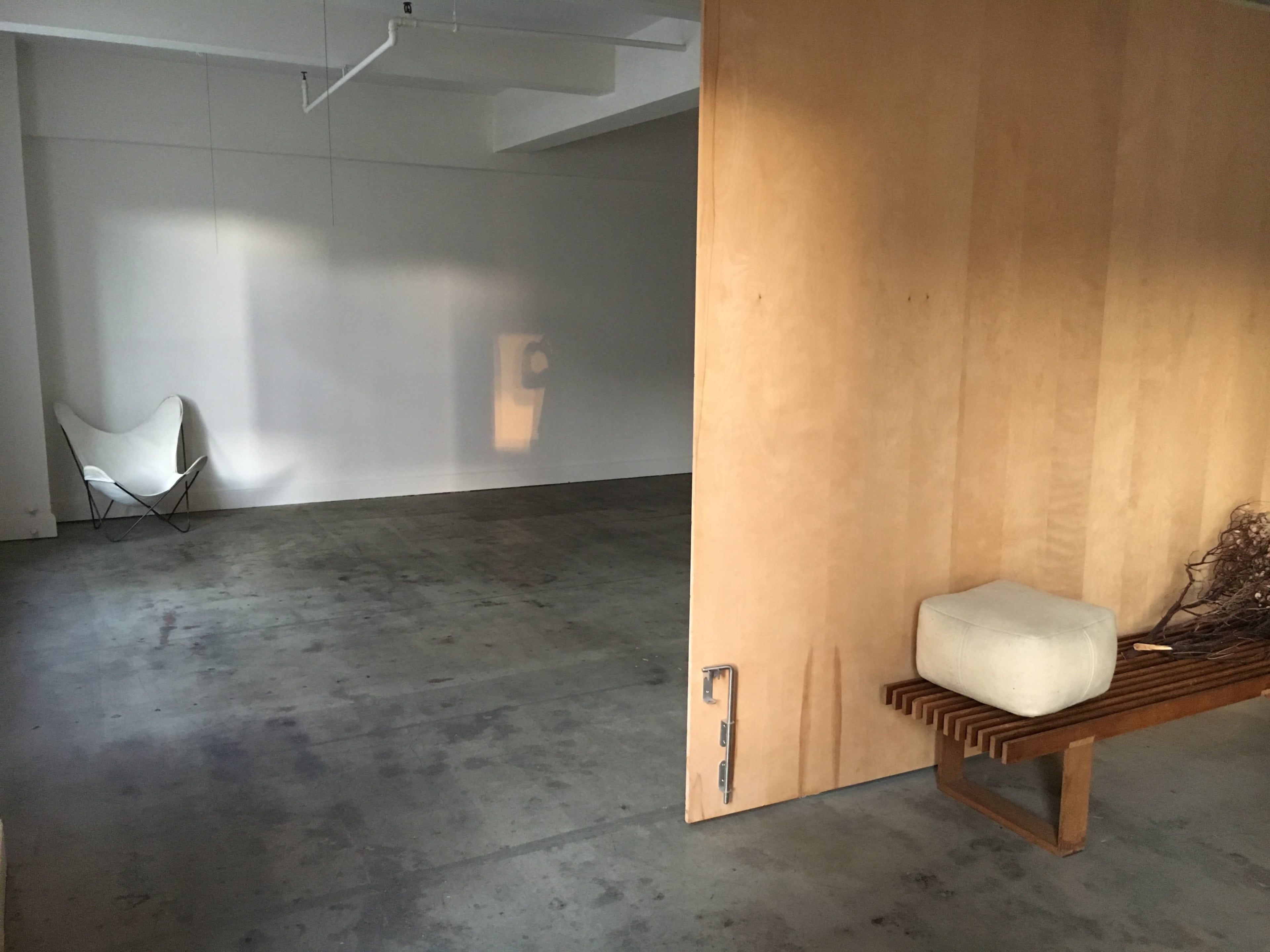 The image shows an interior space with a wooden partition, a light-colored bench, and a decorative object on it, all set against a concrete floor.