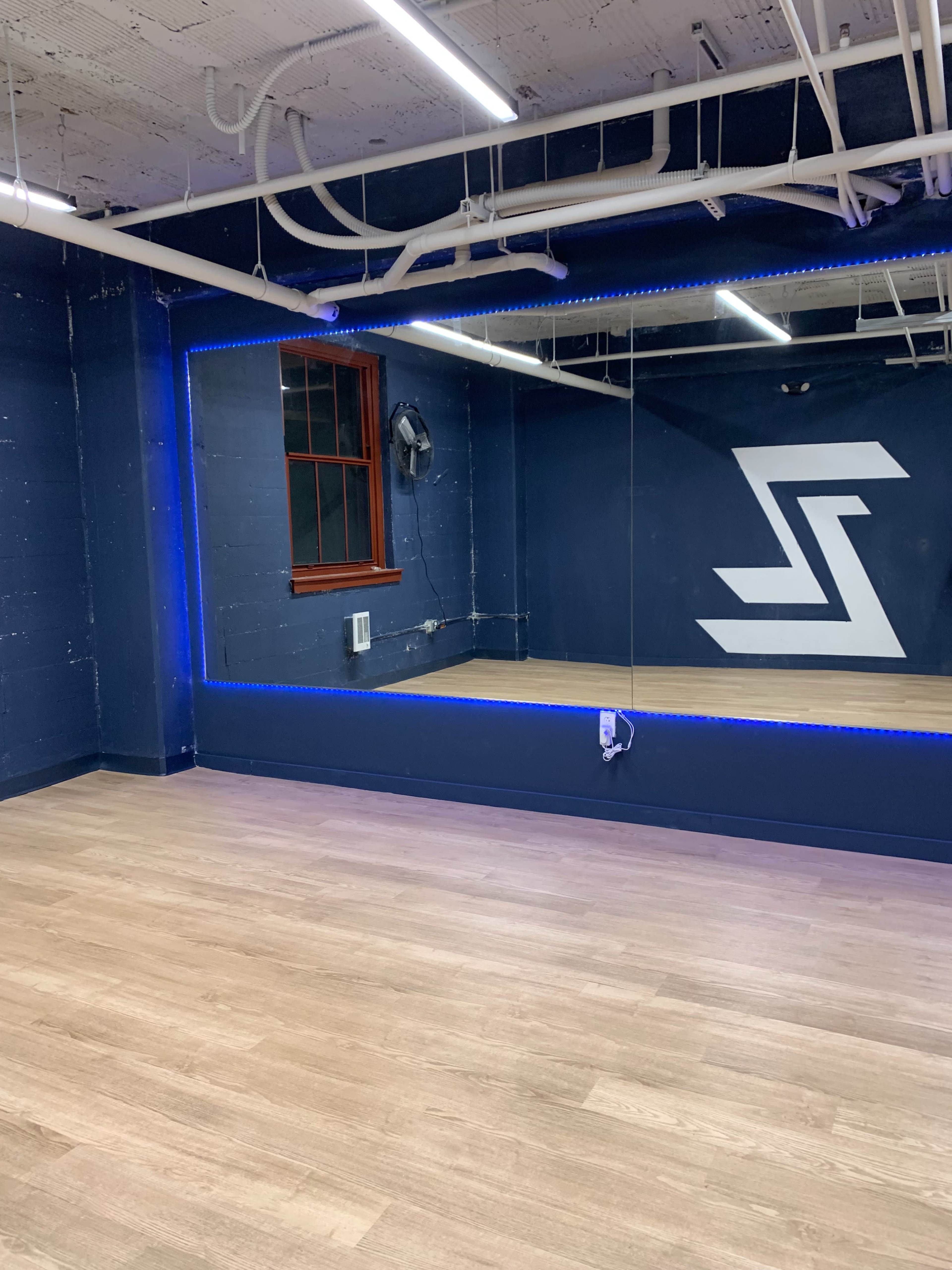 The image shows a dance studio with wooden flooring, a large mirror, blue accent lighting, and a wall featuring a white logo.