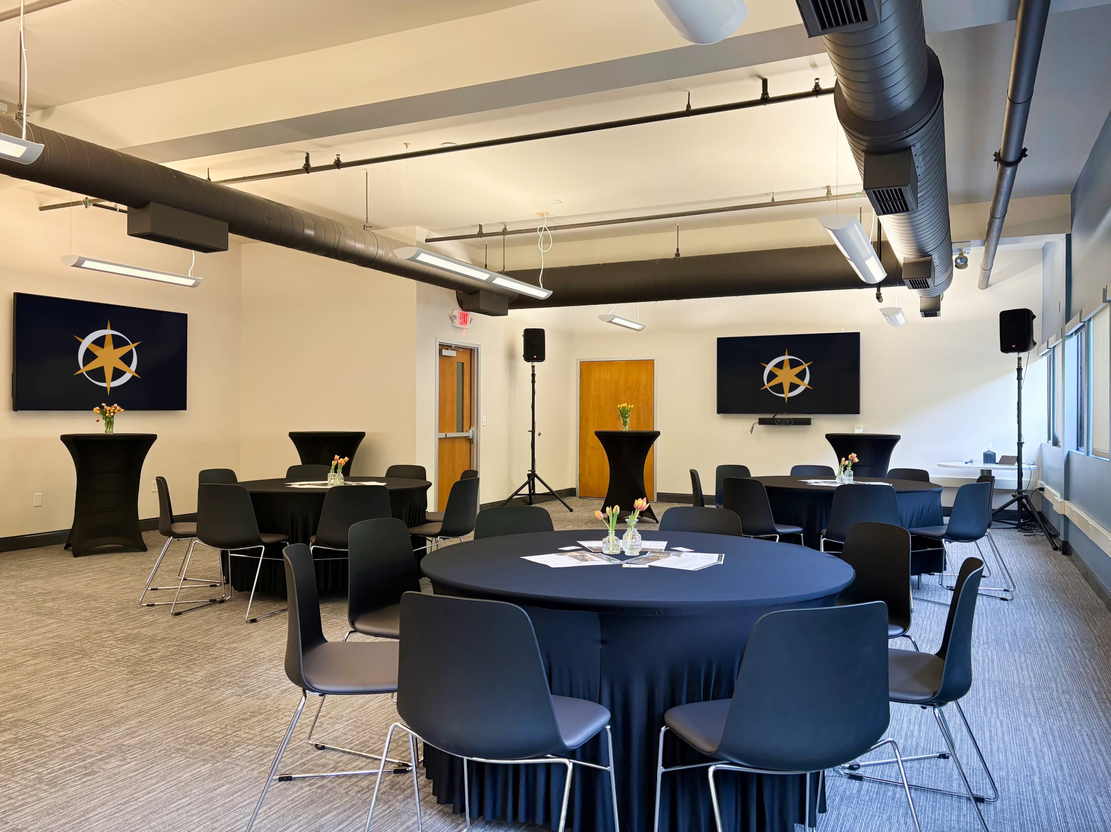 The image shows a conference room arranged with round tables covered in black tablecloths, each set with floral centerpieces, and equipped with large screens displaying a logo on the walls.