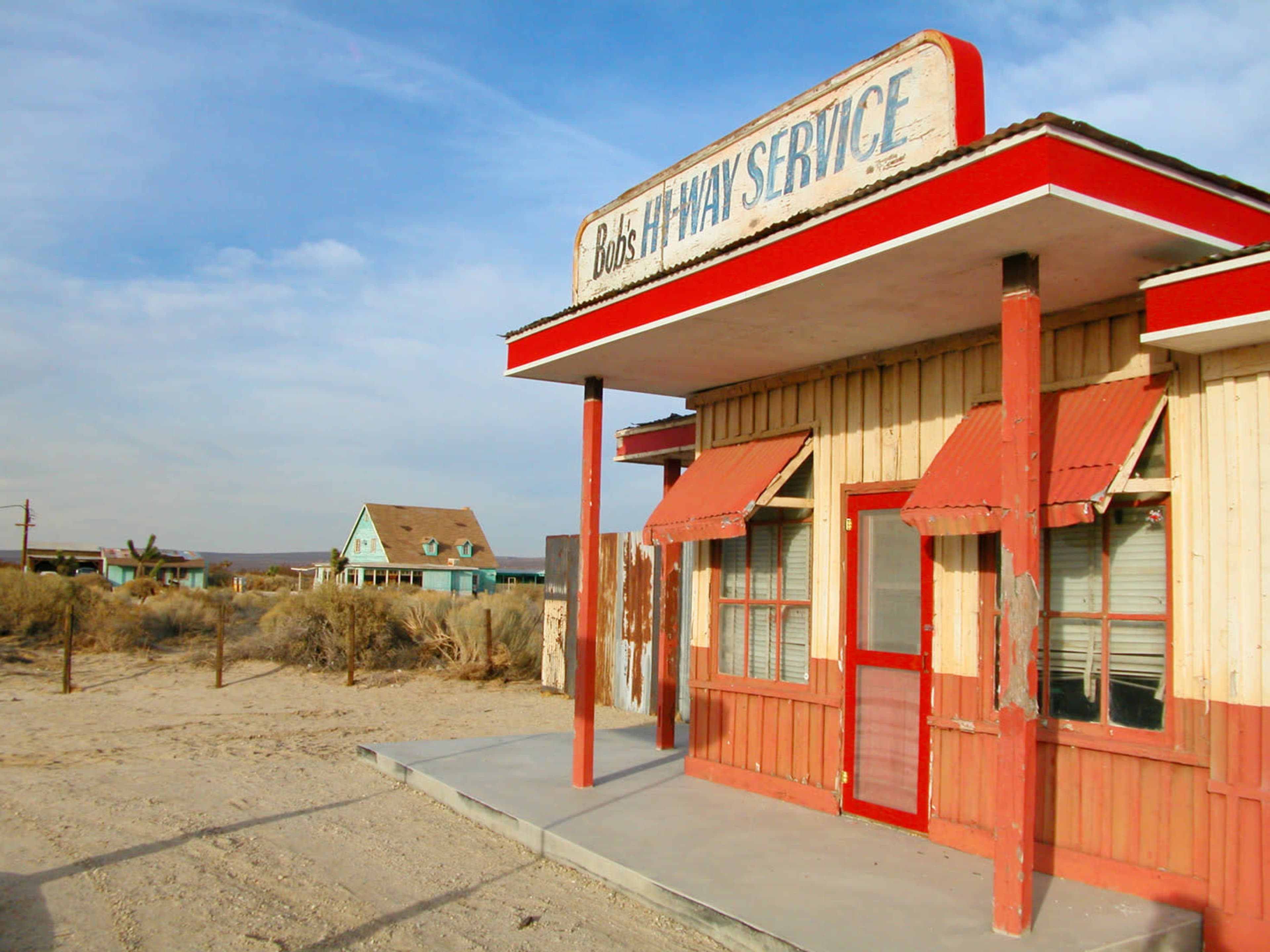 The image shows a weathered service station building with a red trim, labeled "Bob's Highway Service," standing on a sandy lot near an abandoned house.