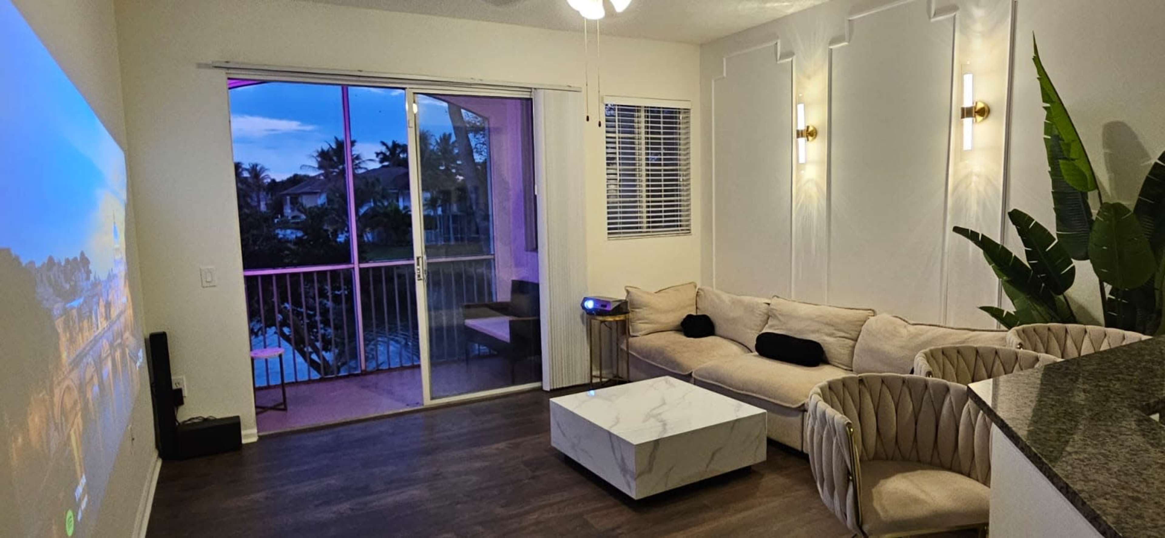 The image shows a modern living room featuring a beige couch, a marble coffee table, and a large window with balcony access, illuminated by wall sconces.