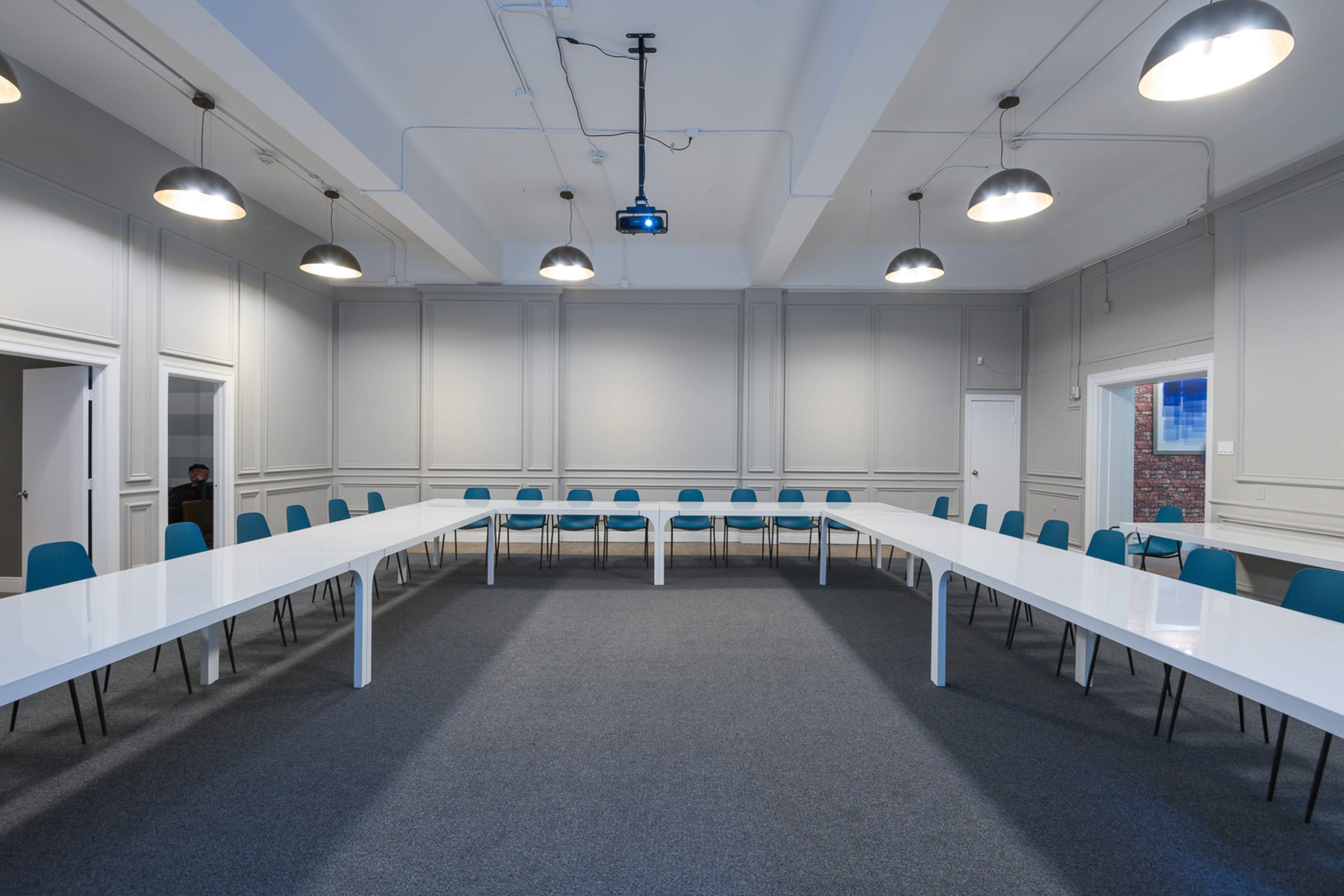 A spacious meeting room features a large white table arranged in a U-shape with blue chairs around it, under industrial-style lighting.