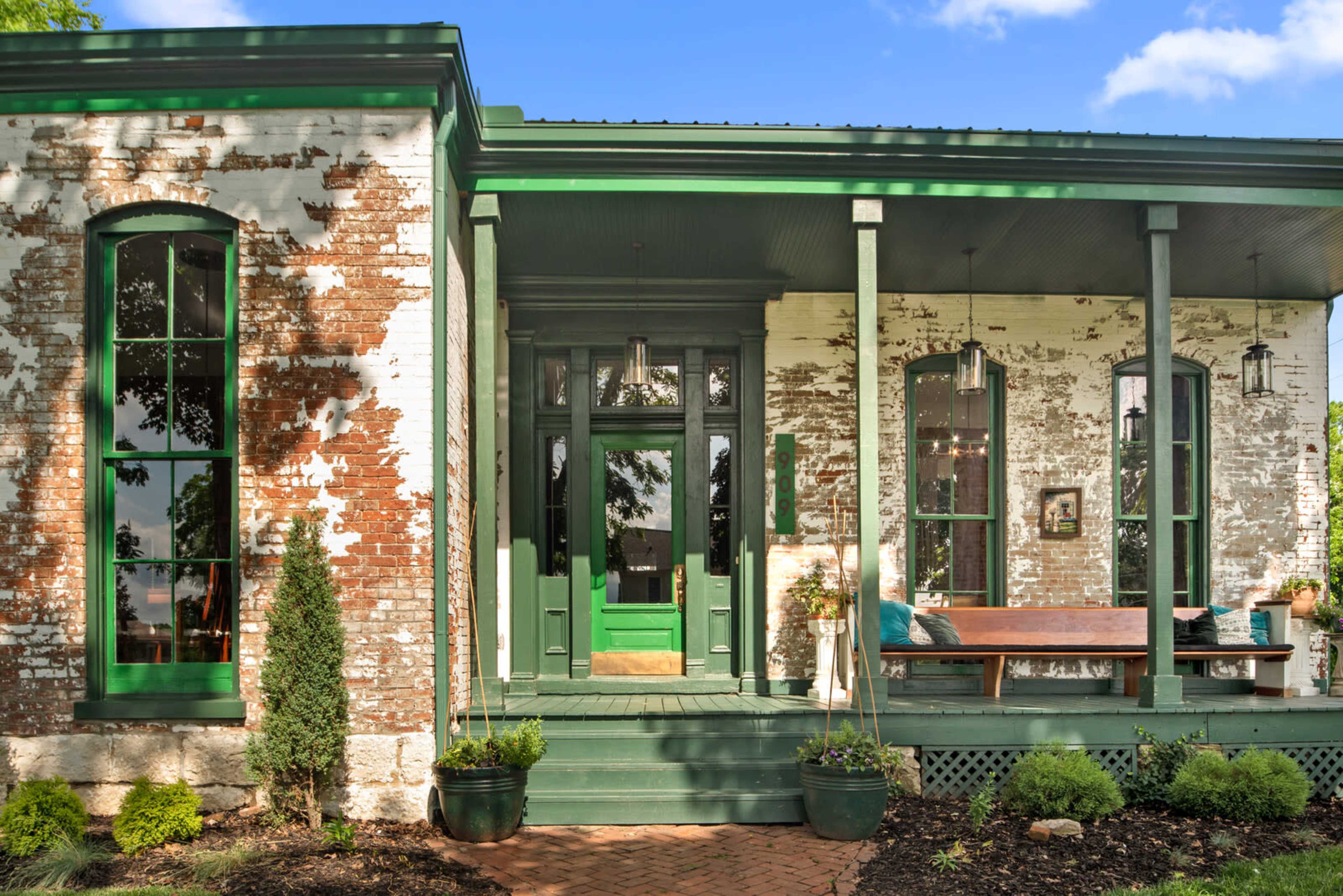A vintage brick house with green trim, featuring large windows and a front porch surrounded by plants and decorative elements.