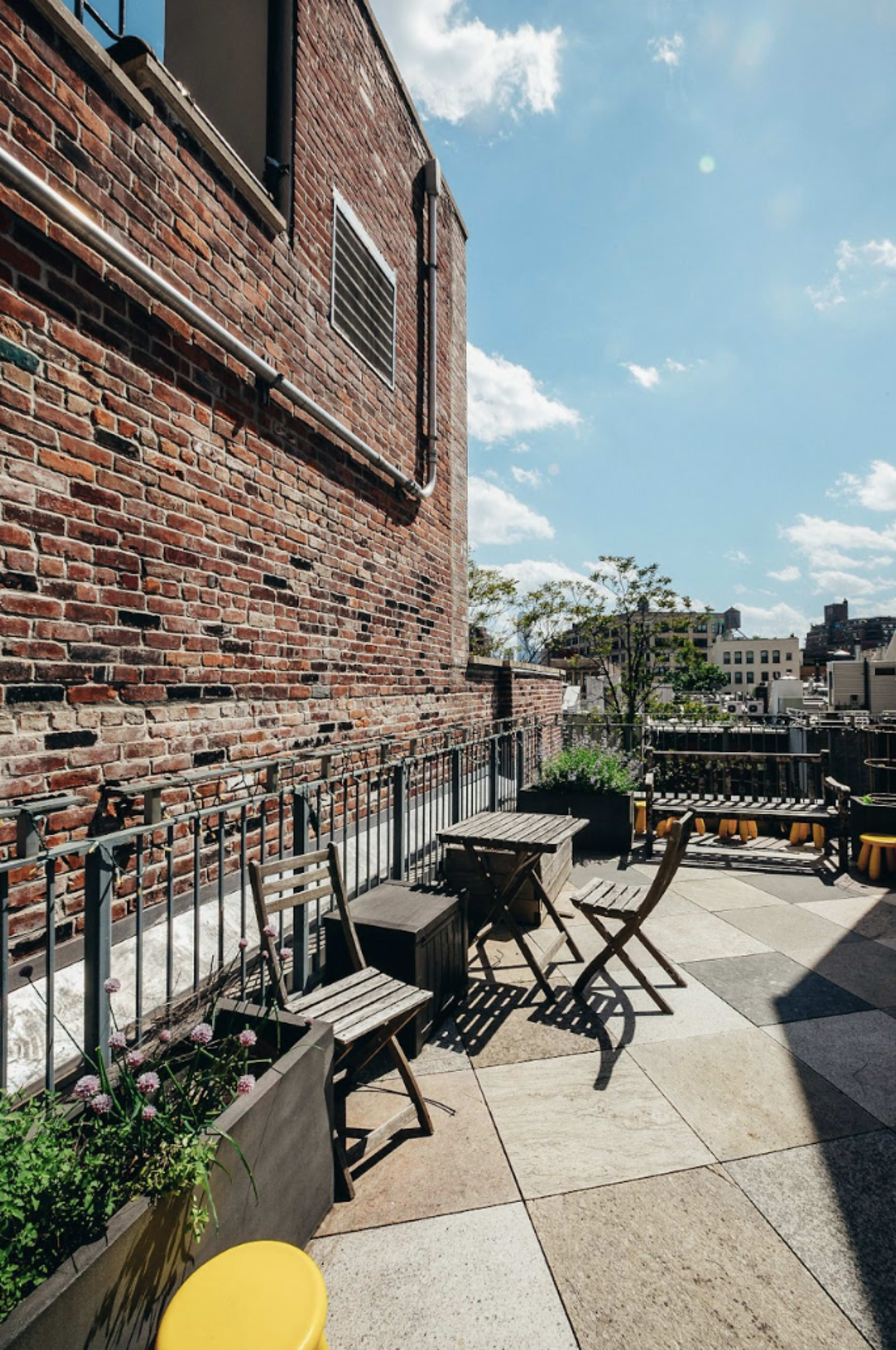 The image shows a rooftop terrace with a brick wall, wooden tables and chairs, and a clear blue sky in the background.