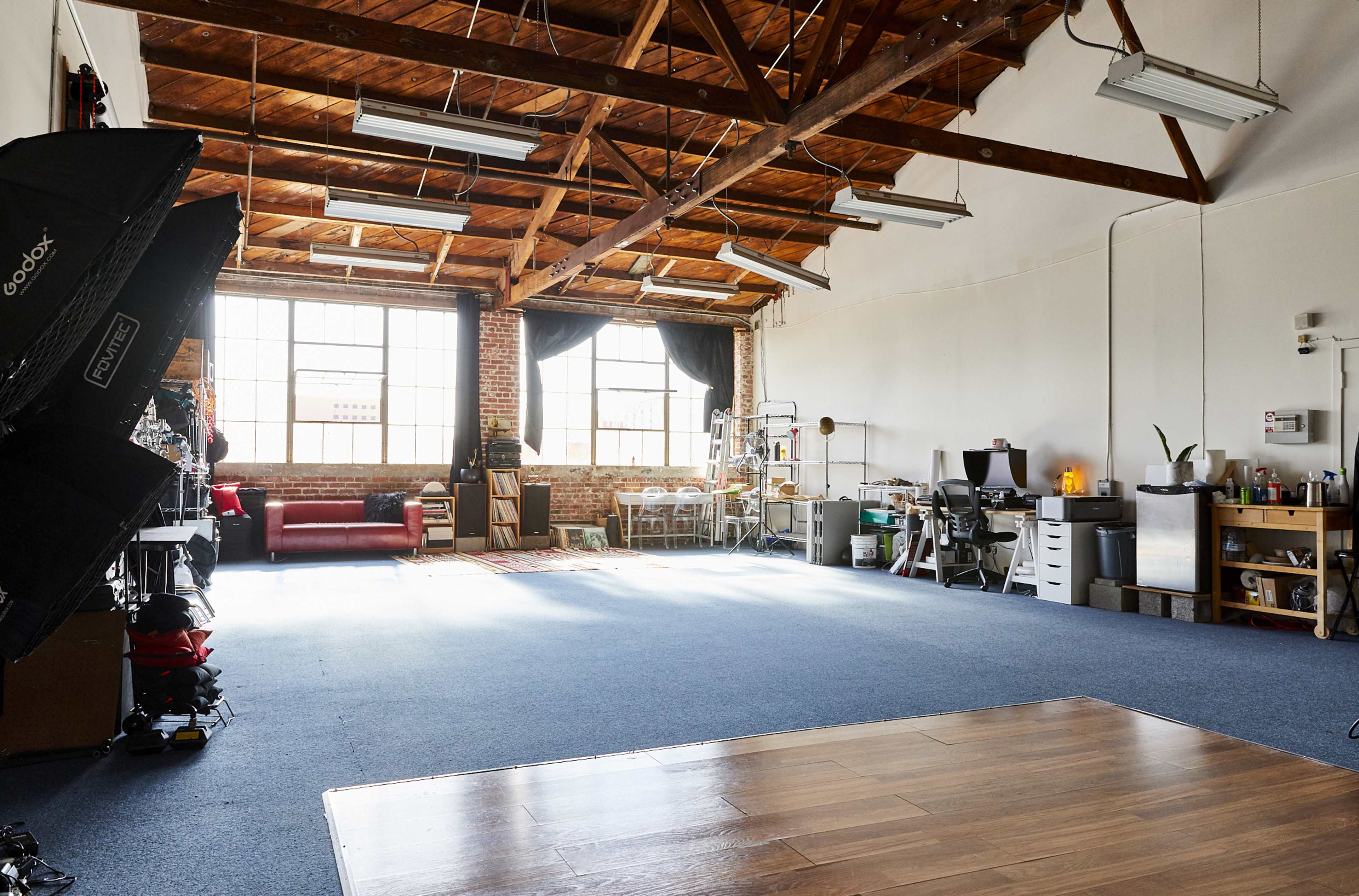The image shows a spacious studio with wooden beams, large windows, and various furniture and equipment arranged around a blue carpeted area.