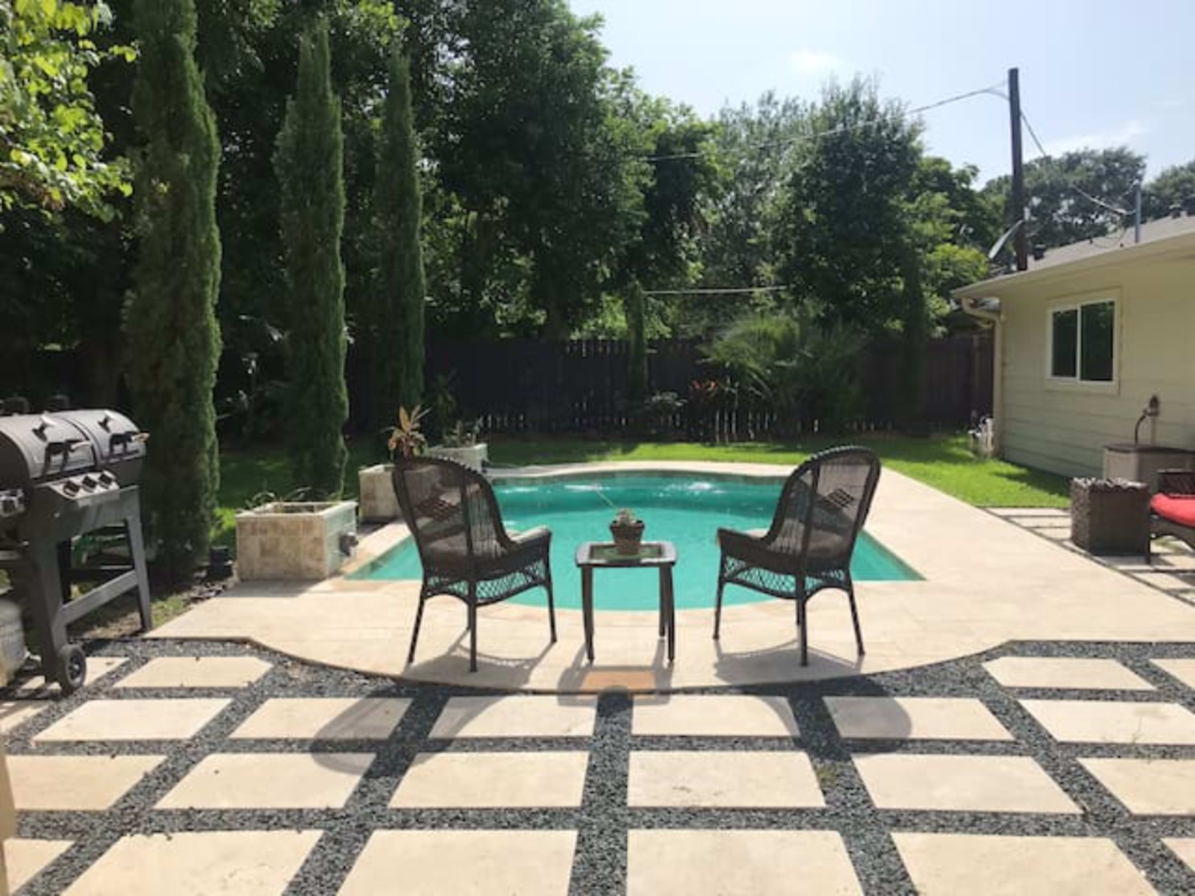 A backyard scene features a swimming pool surrounded by stone pavers, two chairs, a small table, and a grill nearby.