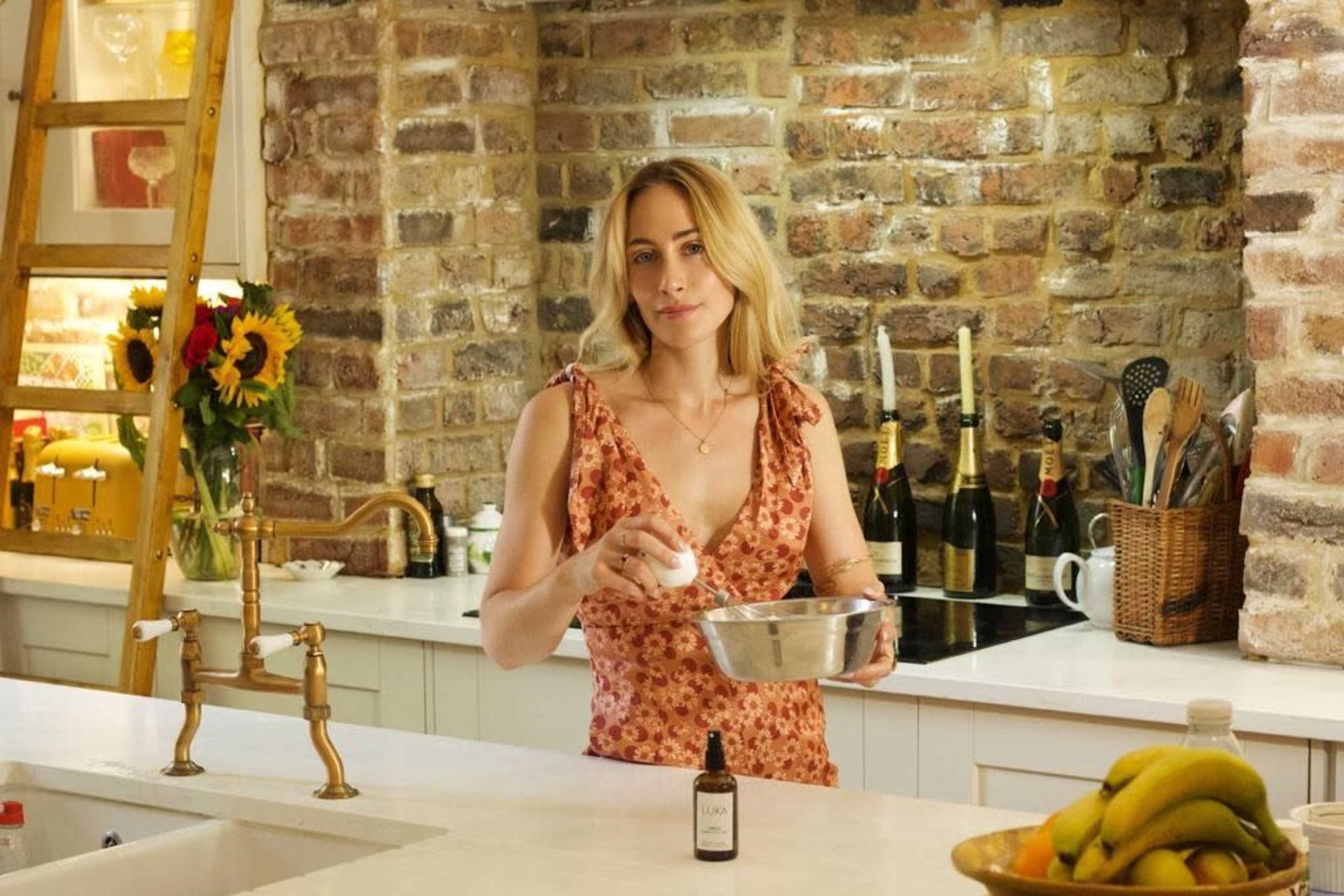 A woman with long hair stands in a kitchen, holding an egg over a metal bowl while surrounded by various kitchenware and a display of fruits.