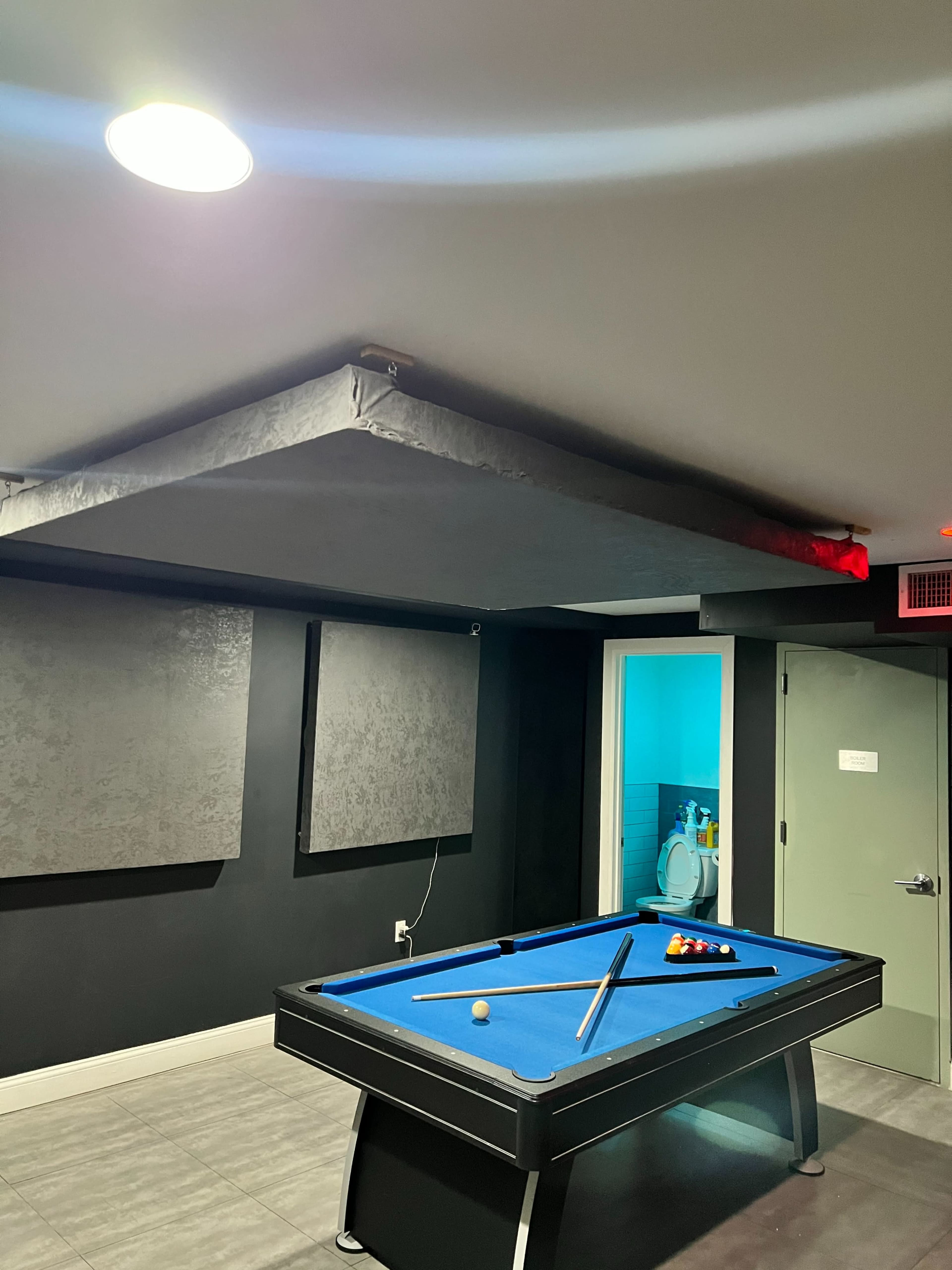 The image shows a room with a blue pool table, grey walls, and acoustic panels, featuring a door that leads to a bathroom.