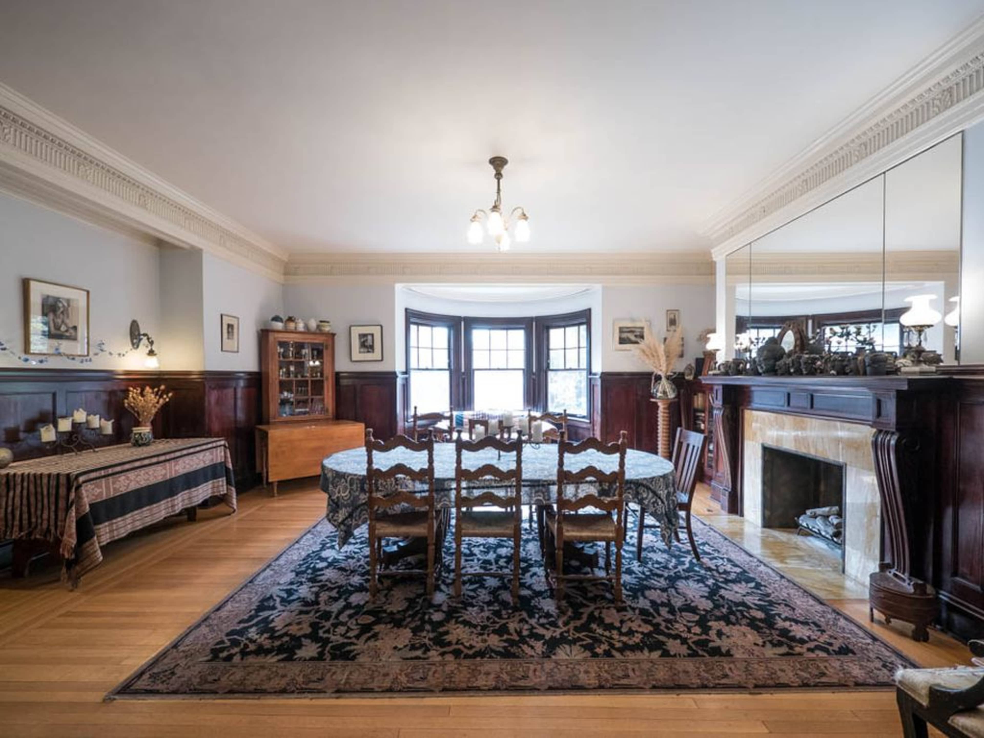 A dining room with a large table surrounded by chairs, a sideboard, and a decorative fireplace, all set within elegant wood paneling and a patterned rug.