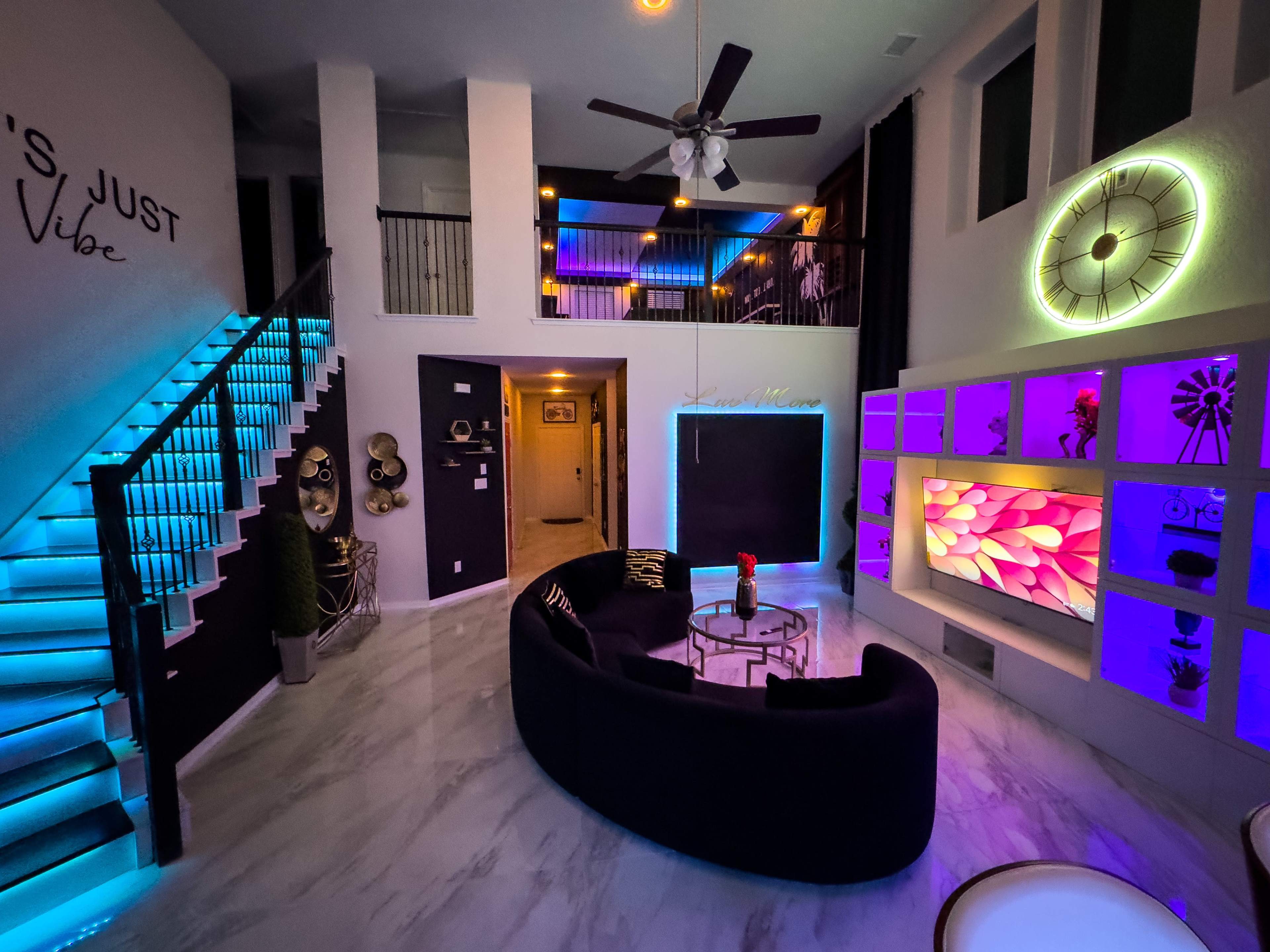 A modern living room with a curved black couch, illuminated stairs, and colorful backlighting on the walls, creating a vibrant atmosphere.