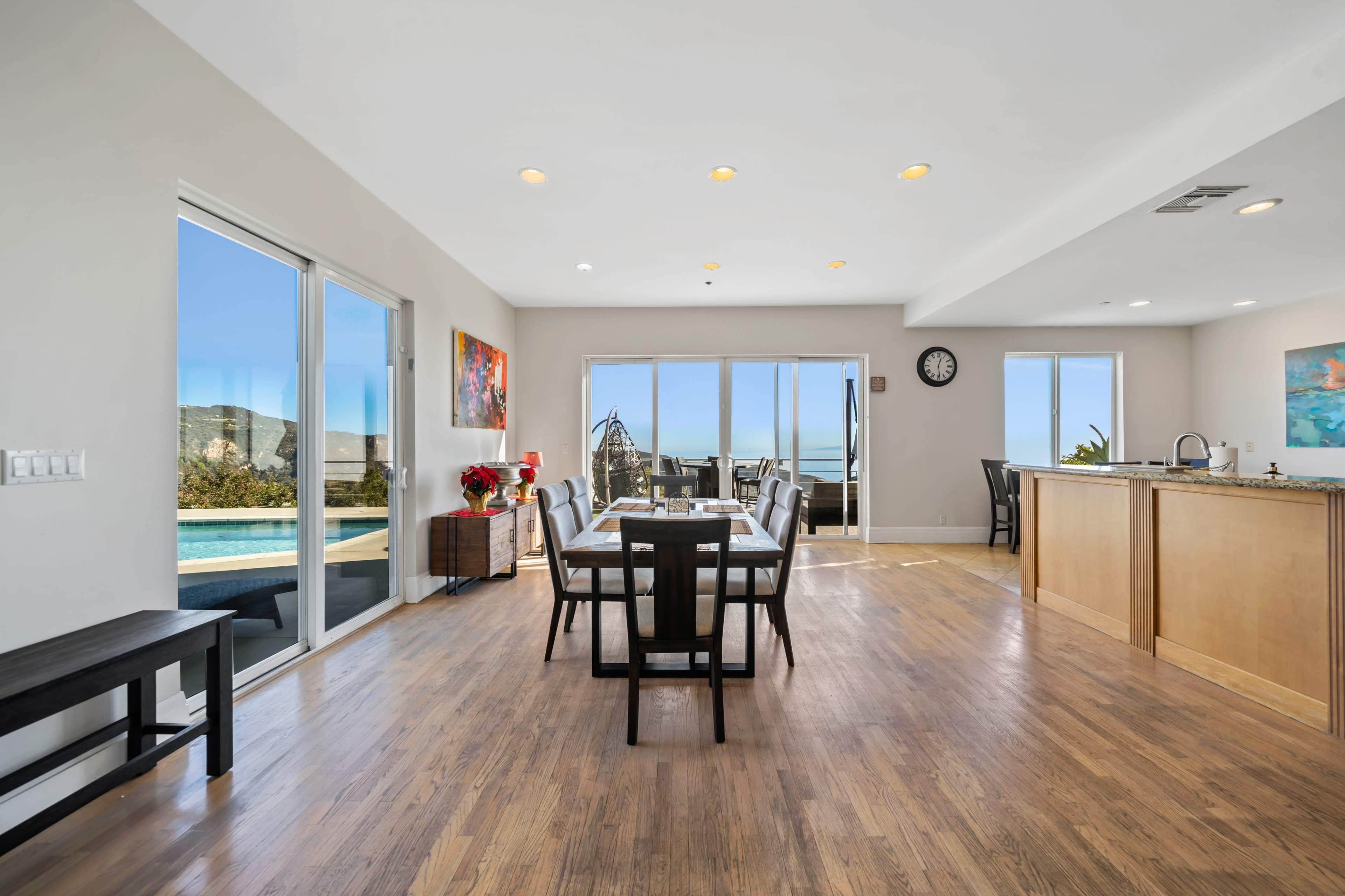 The interior of a modern dining area features a table with chairs, large windows offering a view of the outside, and an open layout connecting to a kitchen.