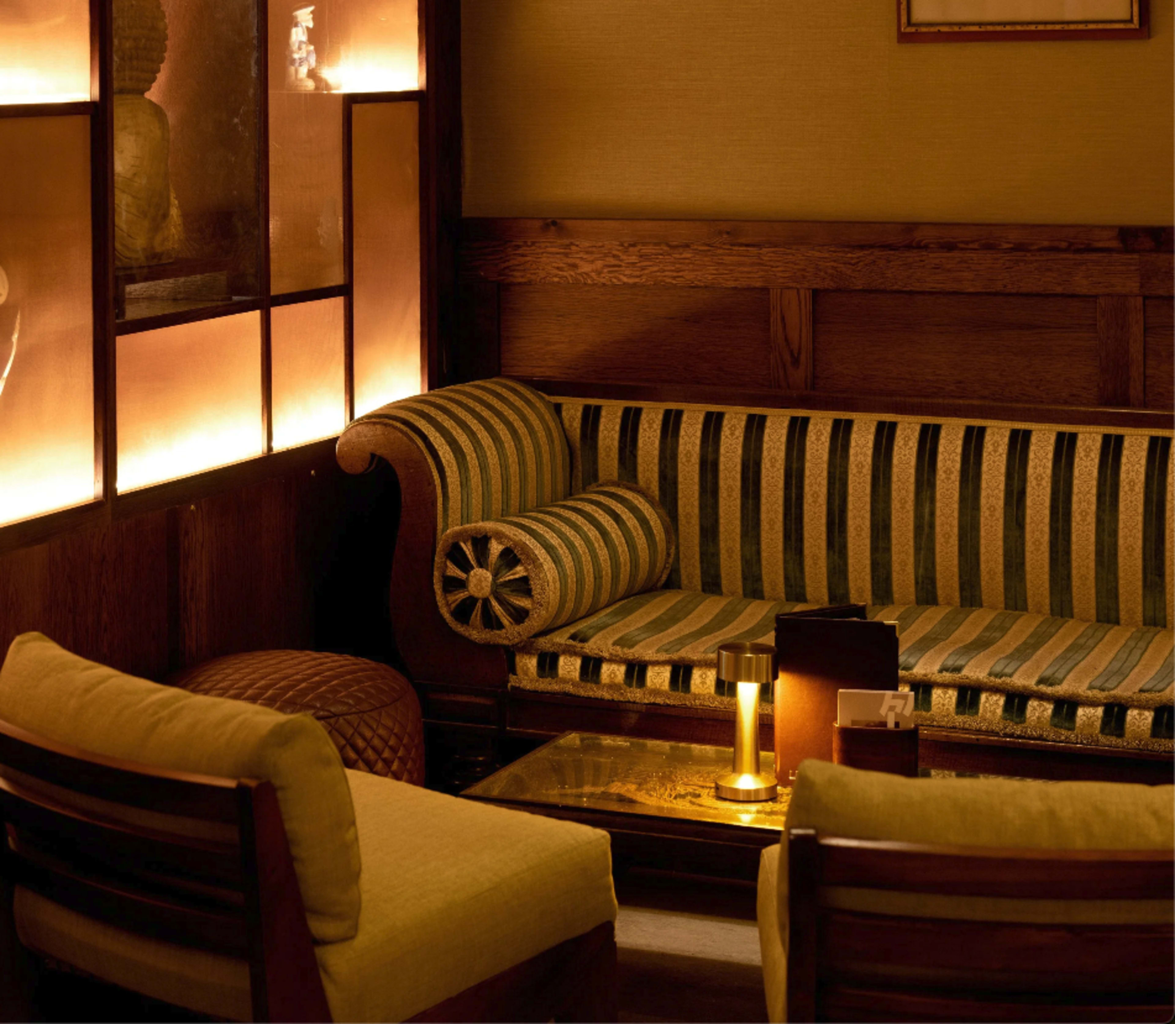 The scene depicts a cozy seating area featuring a striped sofa and two wooden chairs, illuminated by soft lighting.