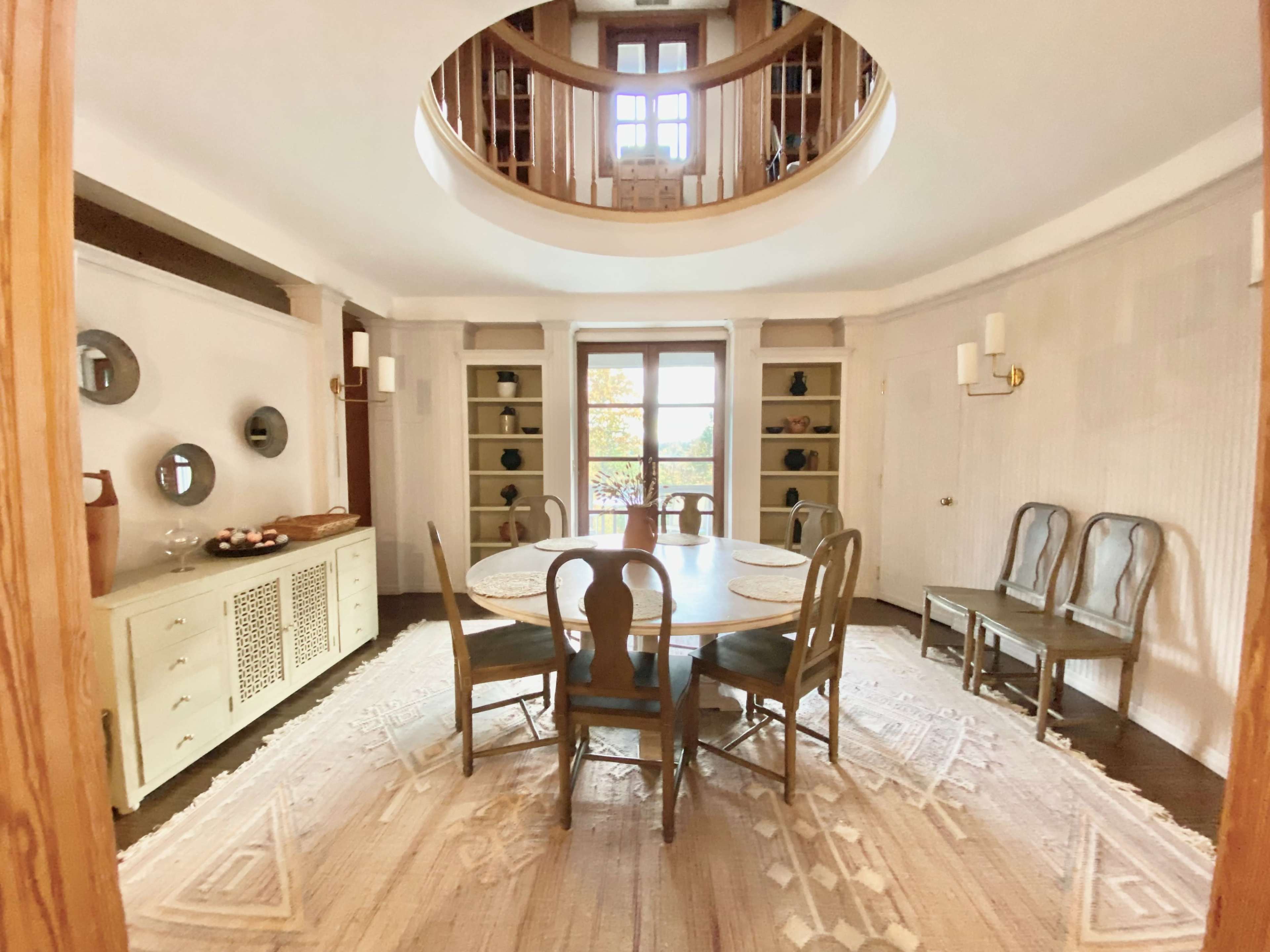 A round dining room with wooden chairs arranged around a table, featuring a circular skylight overhead and built-in shelves against the walls.