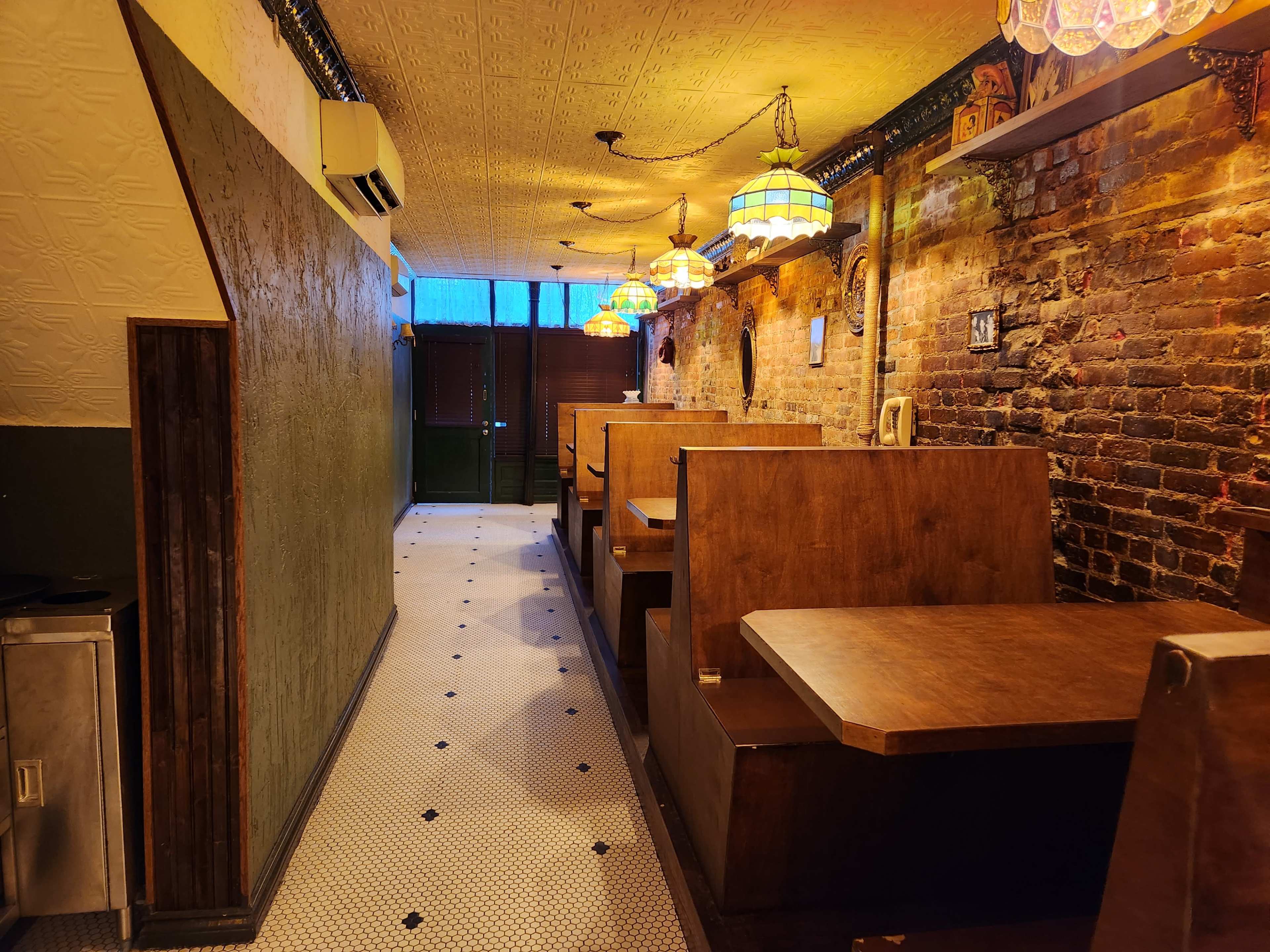 The image depicts a narrow, warmly lit restaurant corridor featuring wooden booths, exposed brick walls, and pendant lighting.