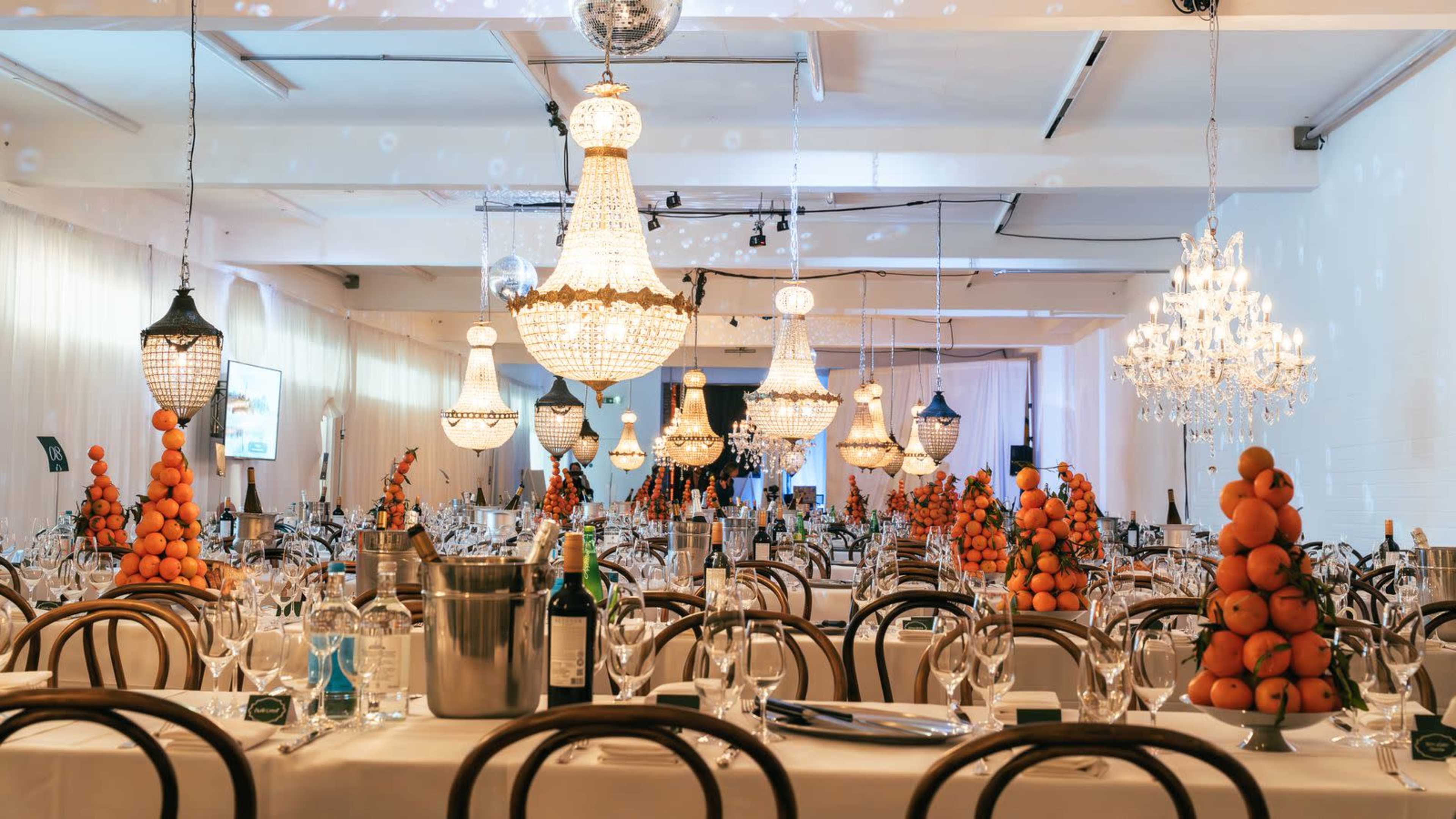 The image shows a well-decorated dining hall with multiple tables set elegantly, each featuring a centerpiece made of stacked oranges and sparkling chandeliers overhead.