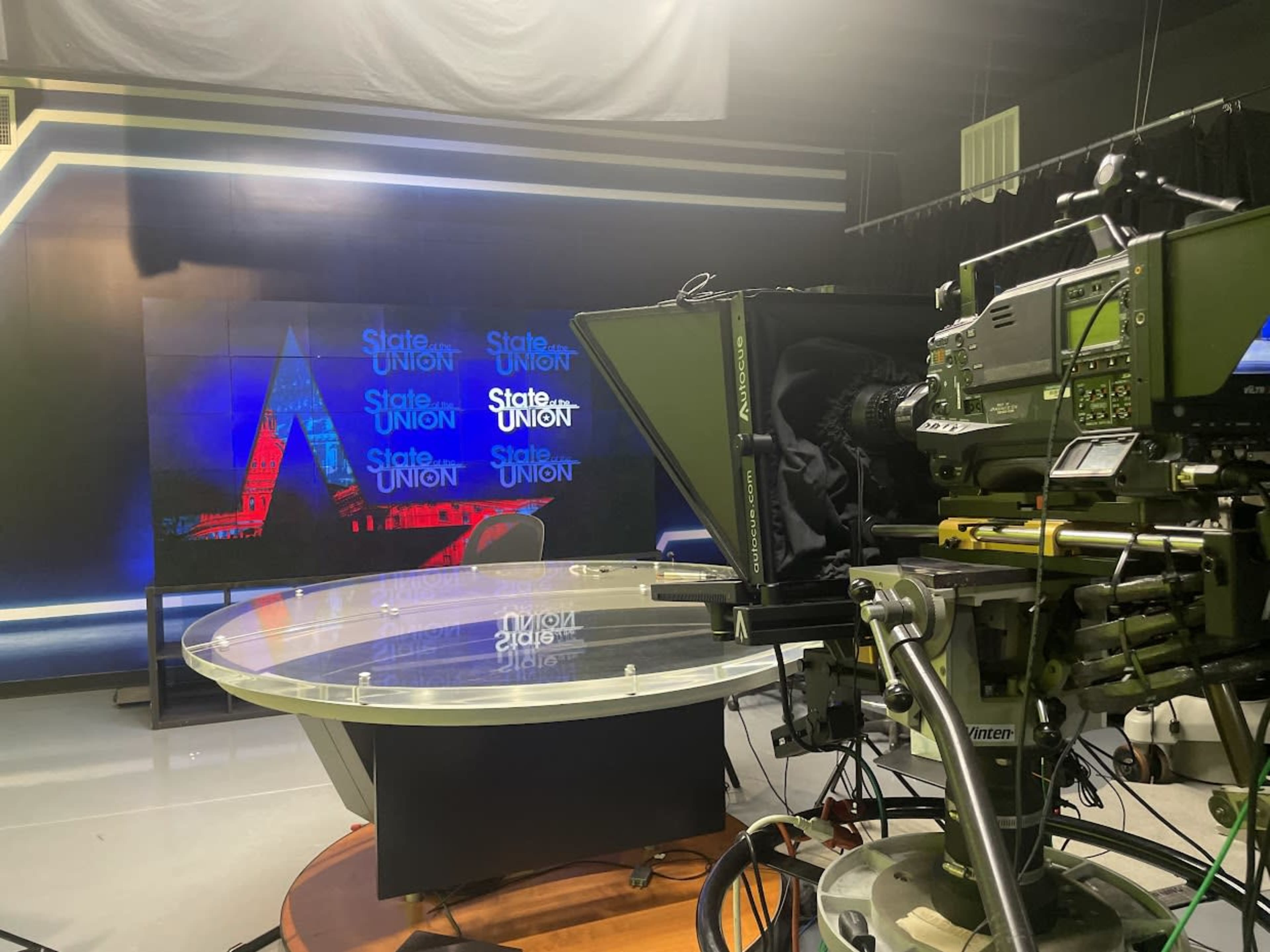 A television news studio features a glass anchor desk, a large background screen displaying "State of the Union," and a professional camera positioned nearby.