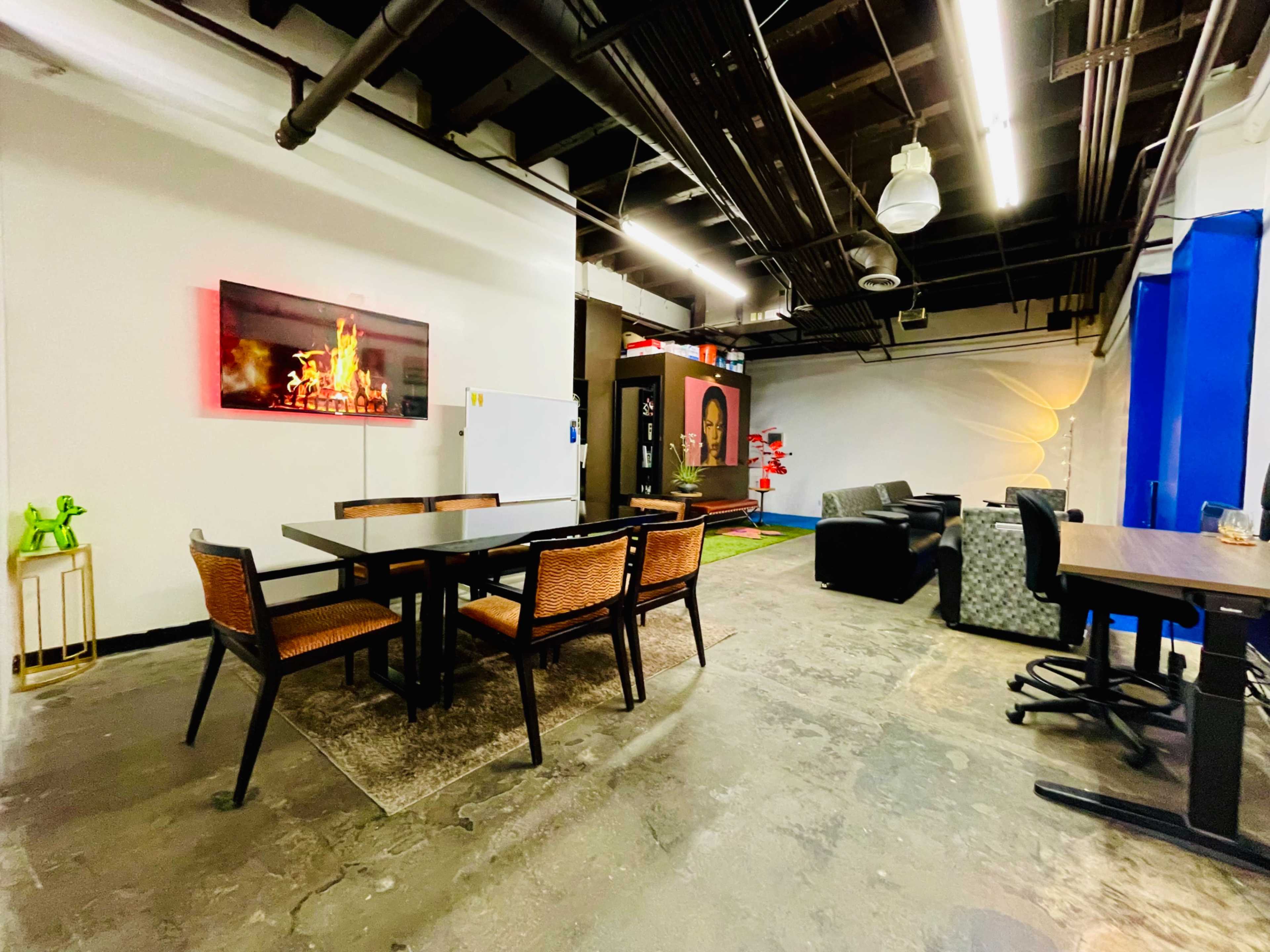 The image shows an open office space featuring a dining table with chairs, a lounge area with sofas, and a small kitchenette.