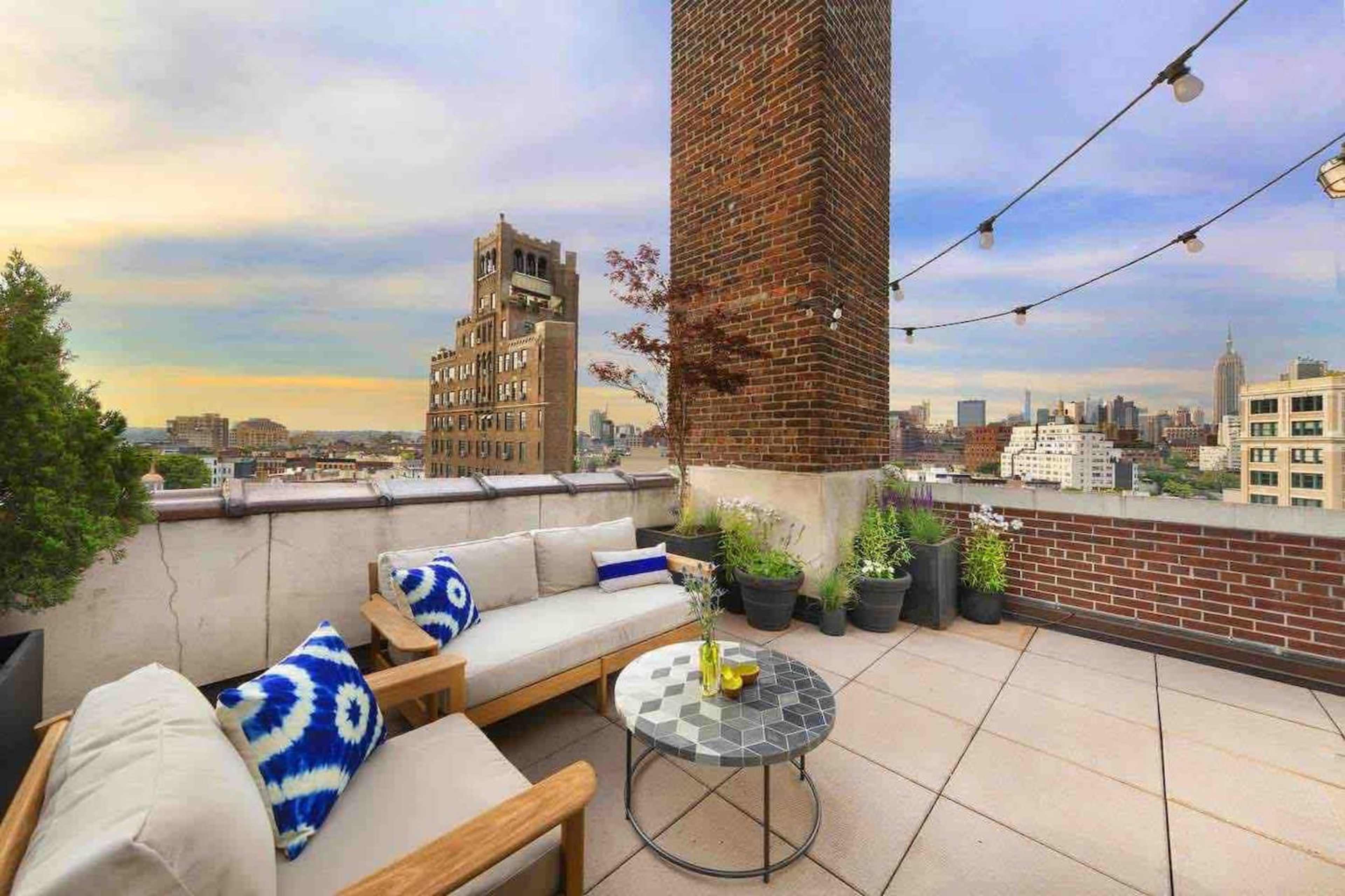 A rooftop terrace features a seating area with a coffee table, surrounded by plants and a view of city buildings.
