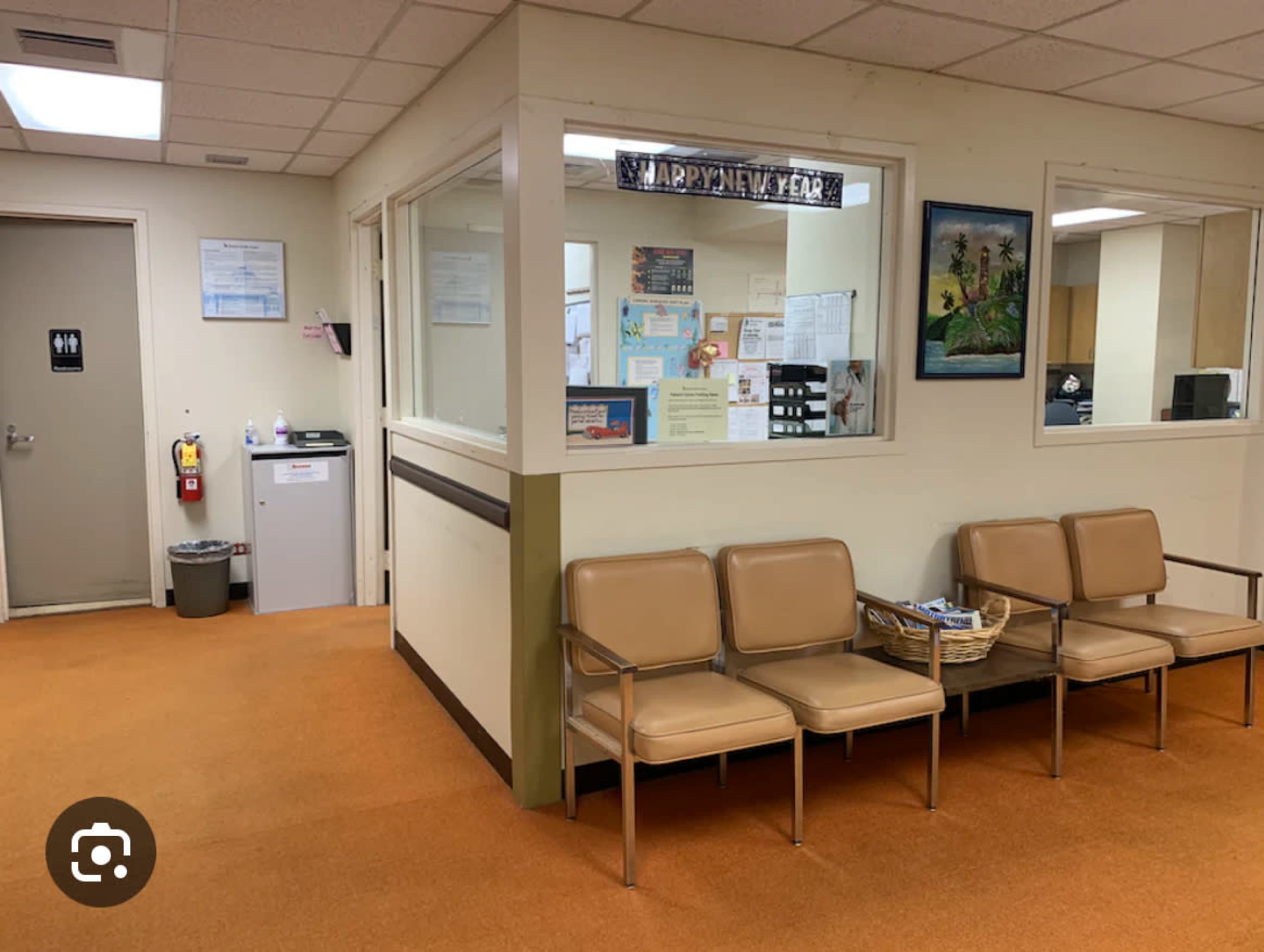 The image shows a waiting room area with orange carpet, three beige chairs, a small table, and an office space visible through a glass partition.