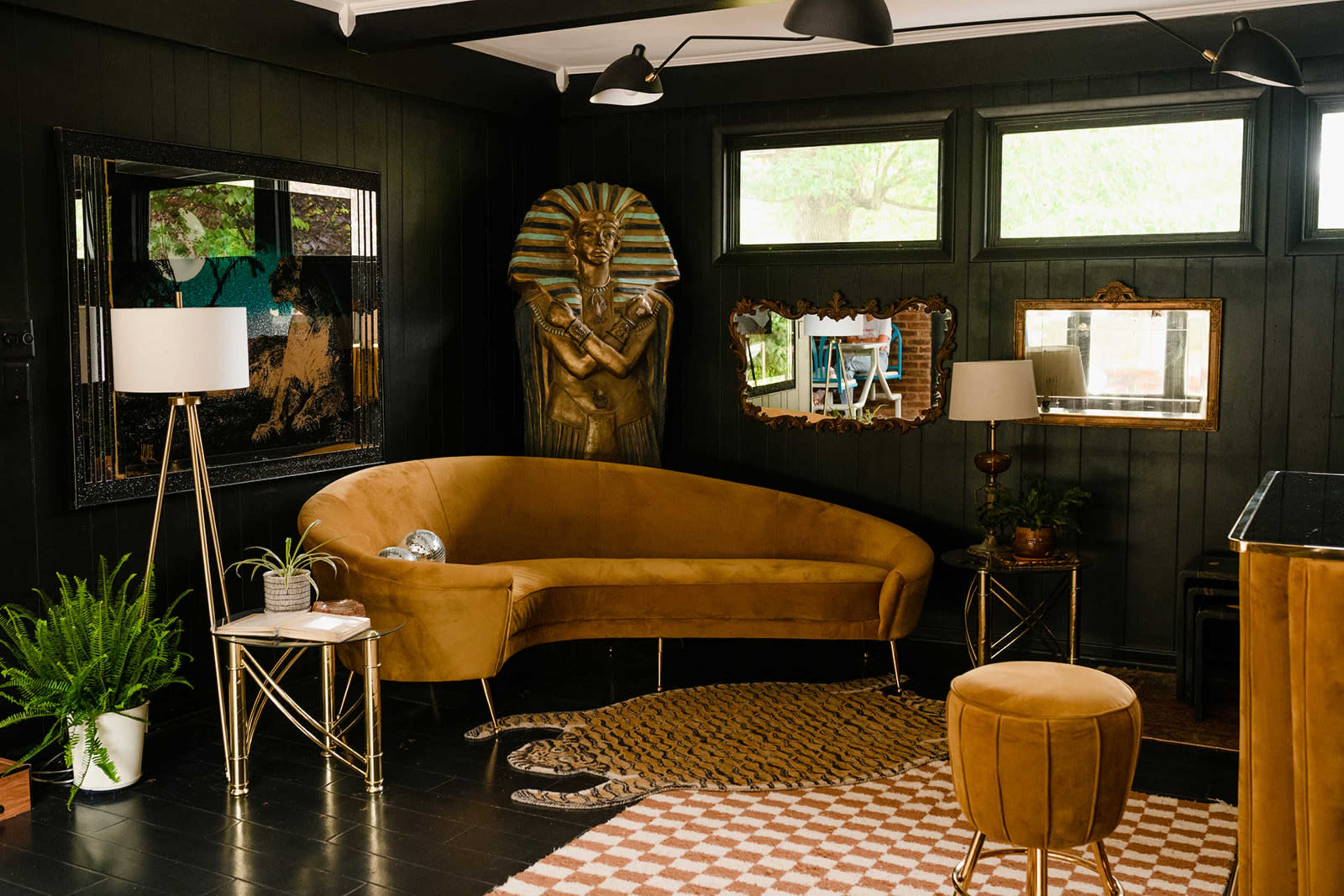 The image depicts a stylish living room featuring a curved brown sofa, gold-accented furniture, and a statue of an Egyptian pharaoh in a dark-paneled space.