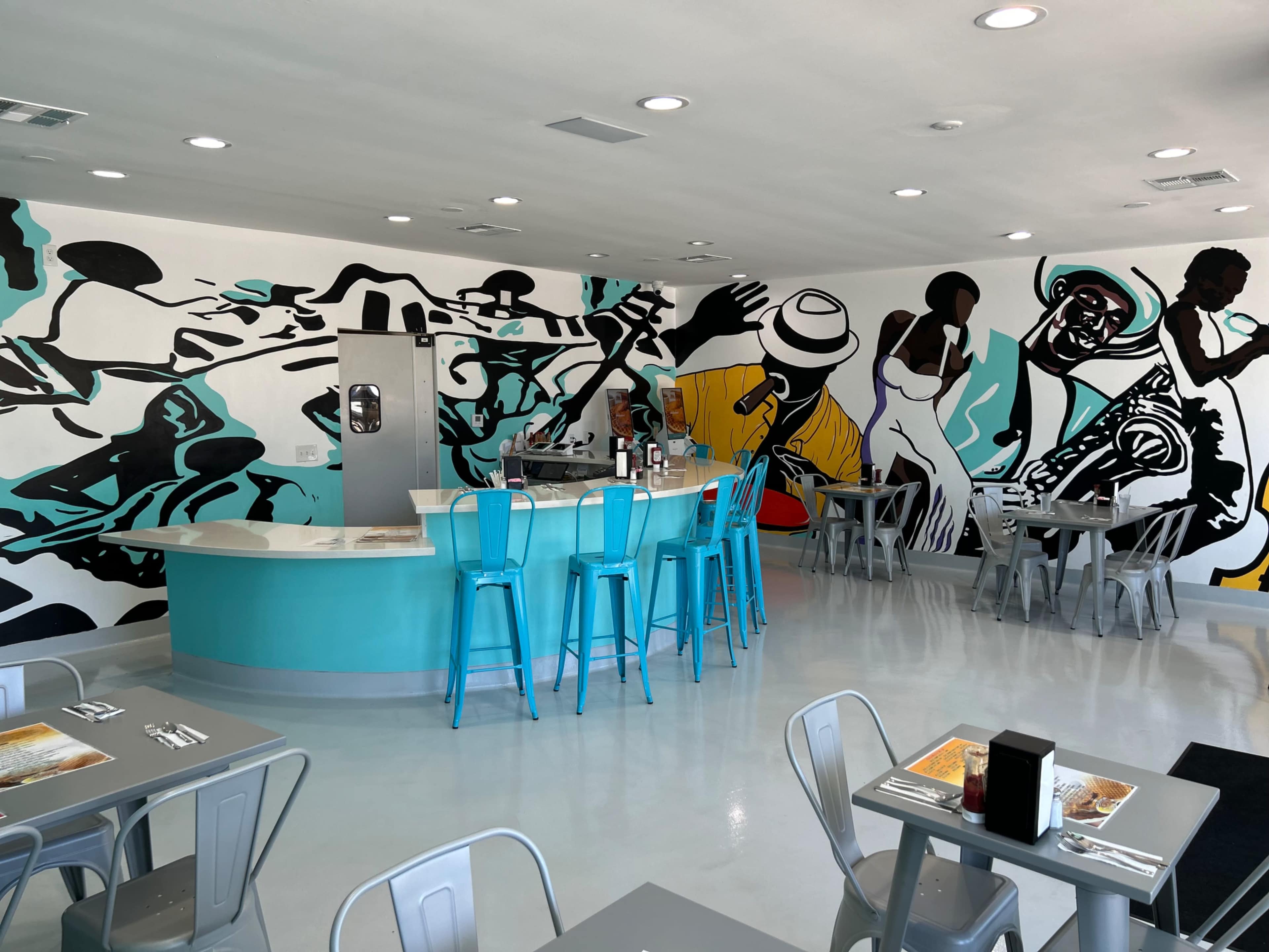 The image shows a brightly colored restaurant interior featuring a central bar with turquoise stools, surrounded by tables and a mural depicting musicians on the walls.