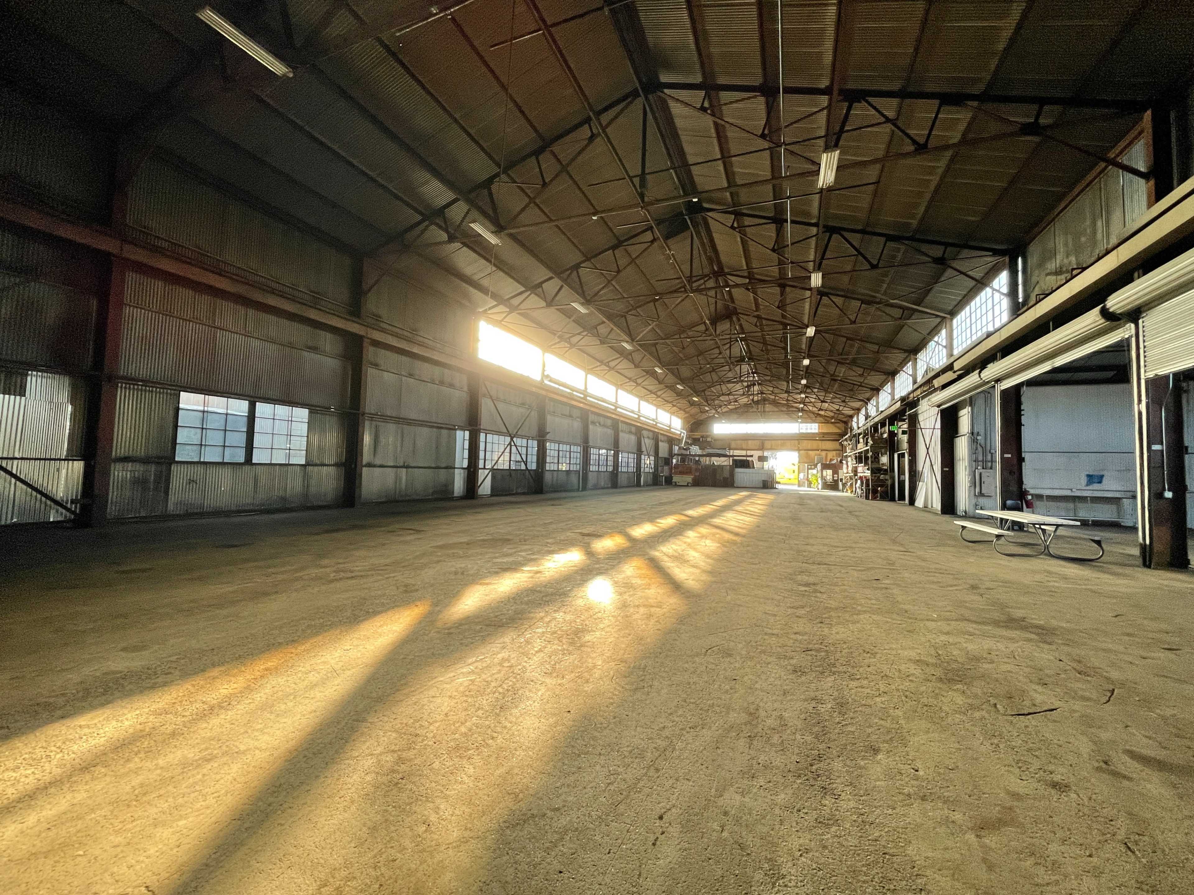 The image shows an empty warehouse with sunlight streaming through the windows, casting long shadows on the floor.