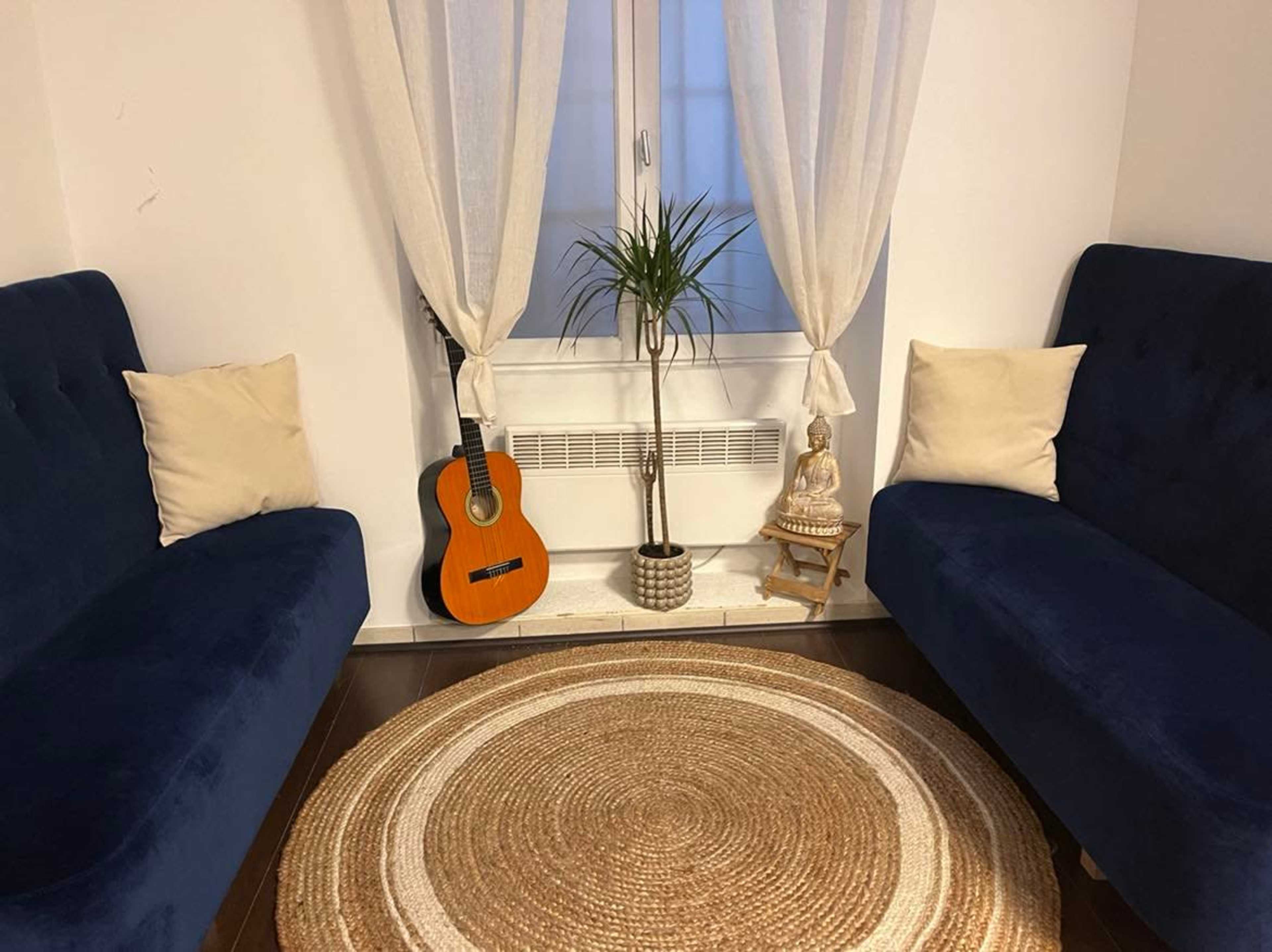 The image shows a cozy corner of a room with two blue sofas facing each other, a guitar propped next to a small statue on a windowsill, and a round woven rug on the floor.