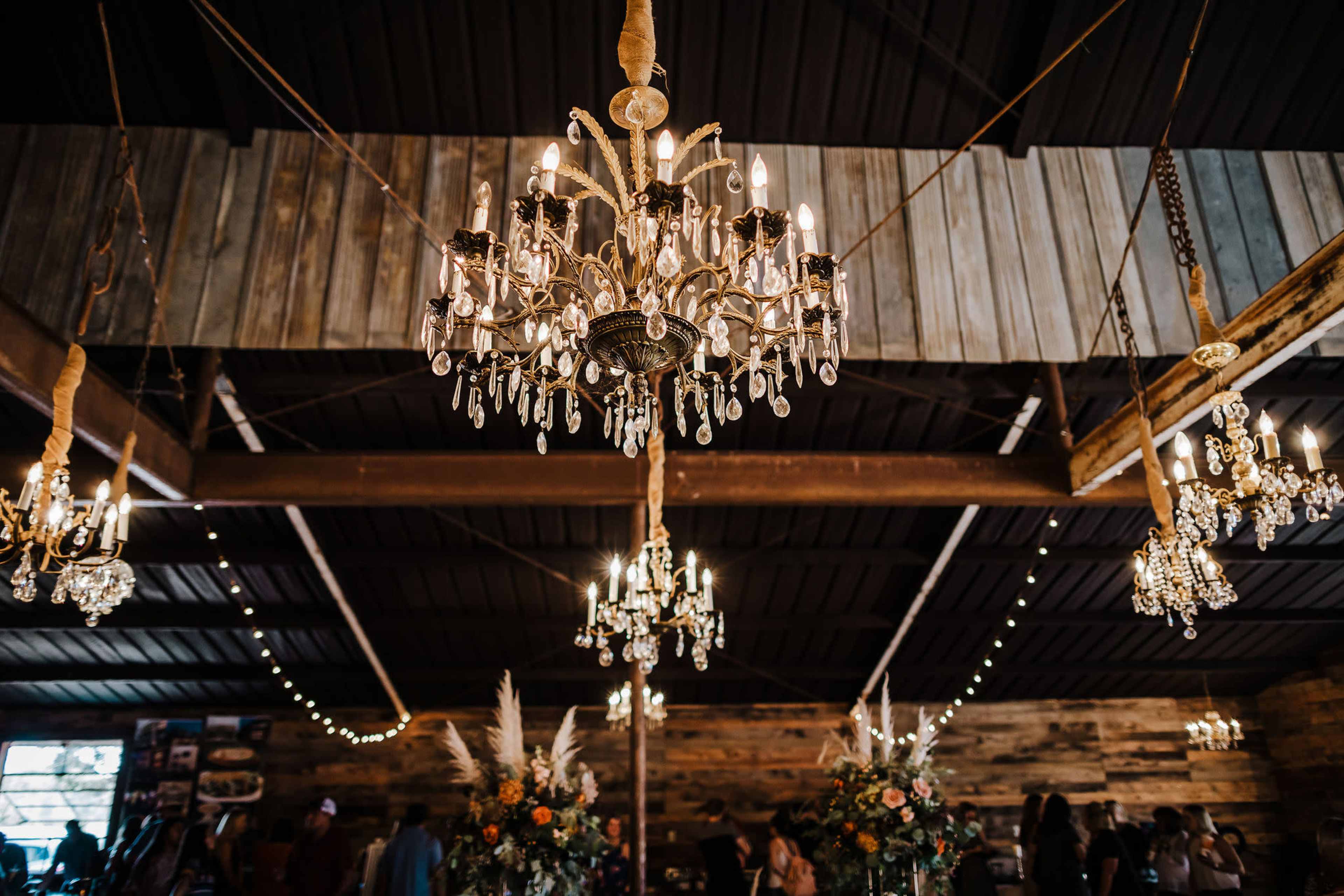 The image shows a rustic interior with multiple ornate chandeliers hanging from a wooden ceiling, adorned with string lights and decorated tables.