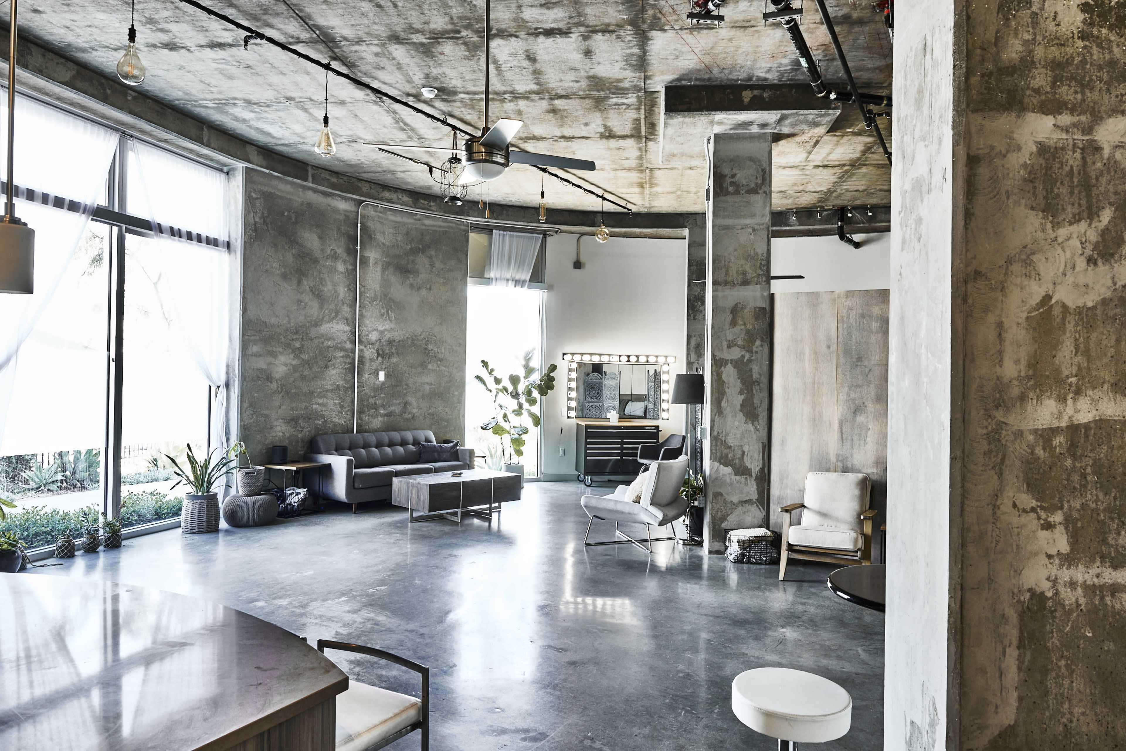 The image shows a spacious, modern living area with concrete walls, large windows, and minimalist furniture.