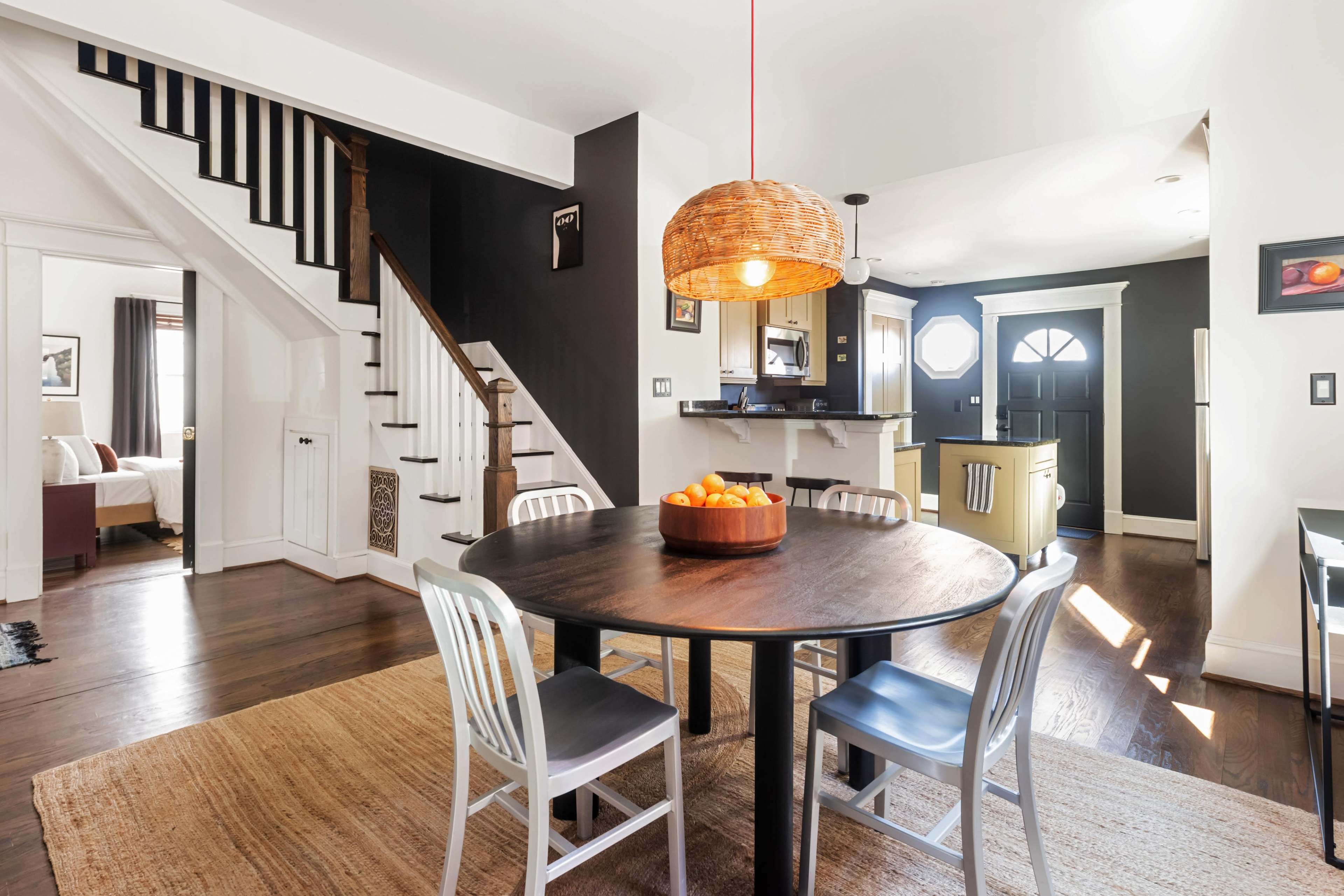 A dining area features a round table with a fruit bowl, surrounded by four chairs, and illuminated by a woven light fixture, with a staircase and kitchen visible in the background.