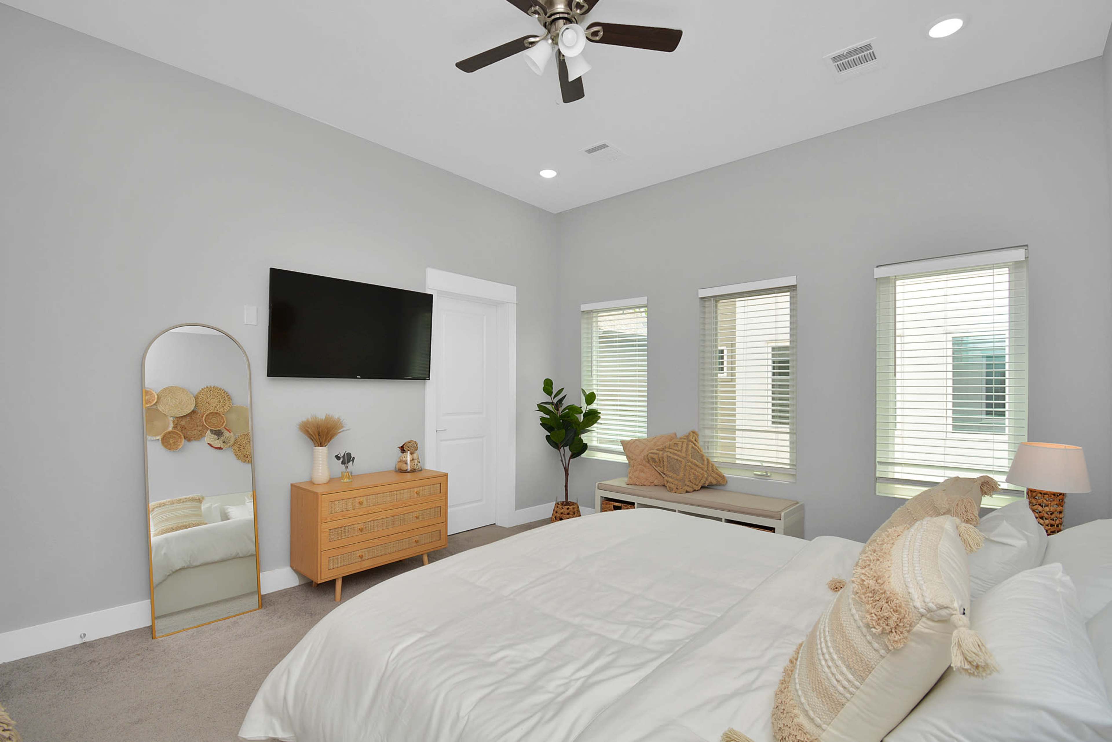 The bedroom features a large bed, a wooden dresser, a wall-mounted TV, and two windows with blinds, creating a bright and functional space.