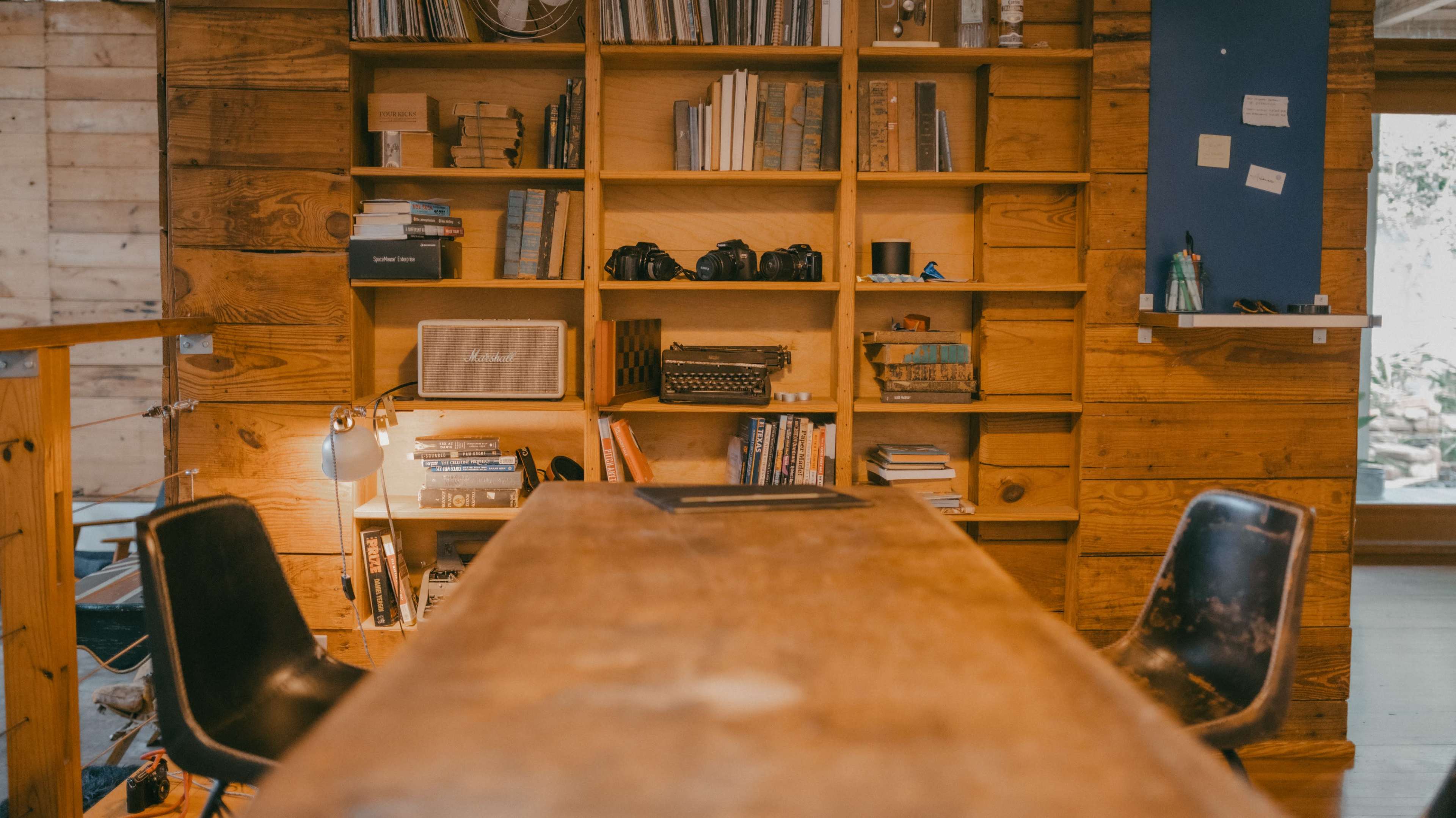 The image shows a wooden table in a cozy room with a bookshelf filled with various items, including books and cameras, and two black chairs positioned at the table.