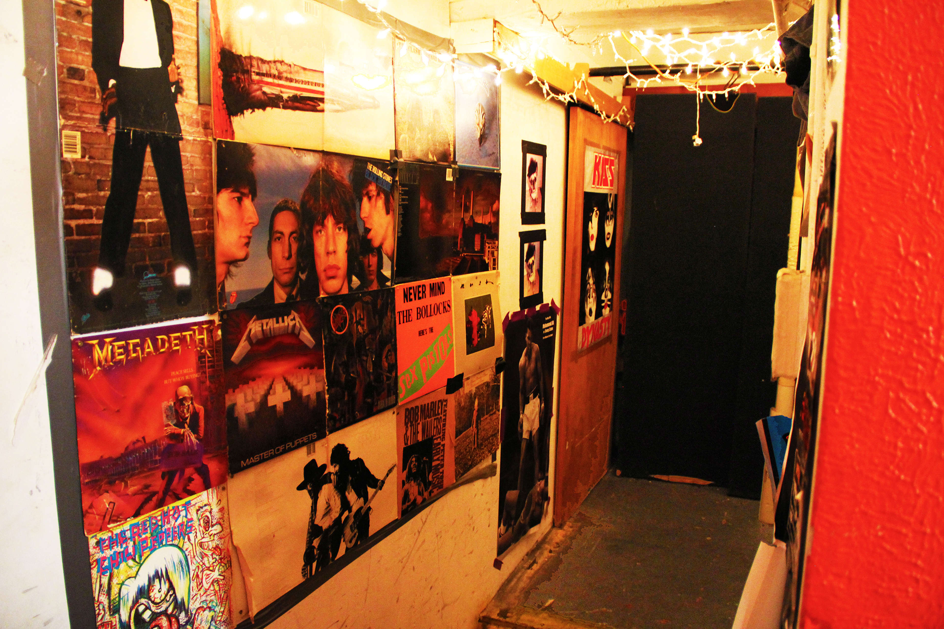 A narrow hallway adorned with various rock music album covers on the walls, illuminated by string lights overhead.