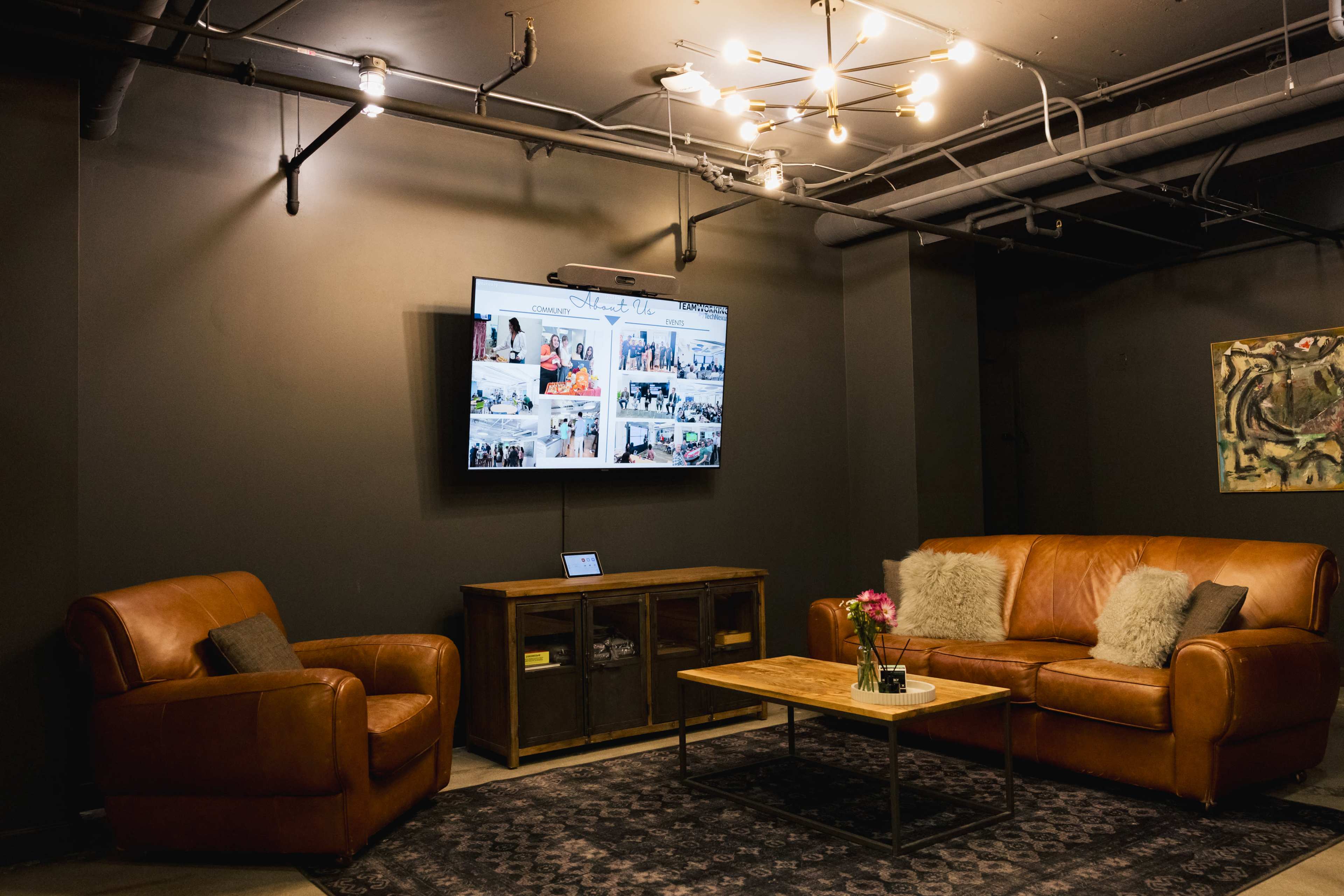 The image shows a cozy lounge area with a brown leather sofa and armchair, a wooden coffee table, and a television mounted on the wall displaying a collage of photos.