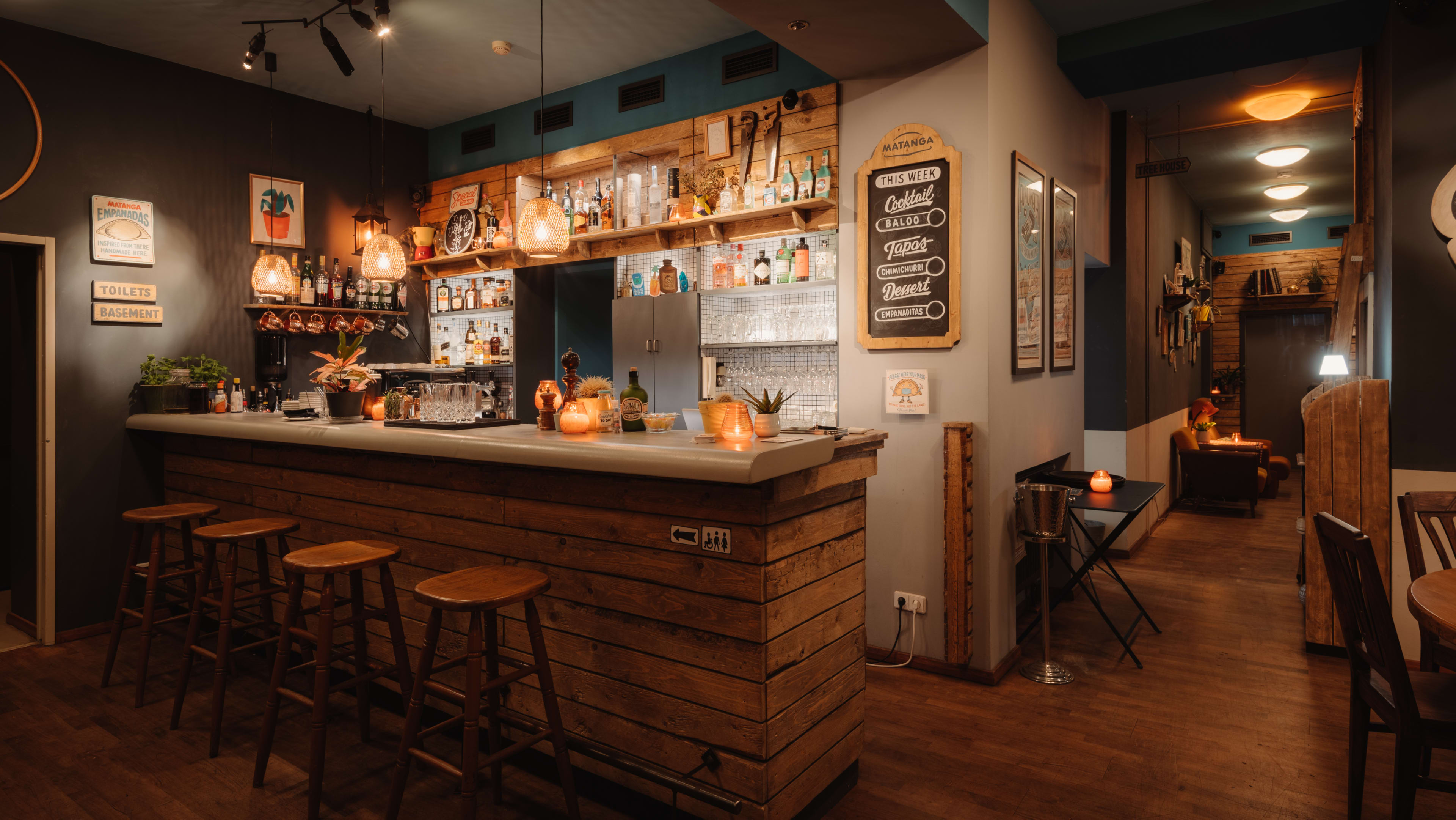 A rustic bar area with wooden stools, a variety of bottles on shelves, and softly glowing candles on the counter.