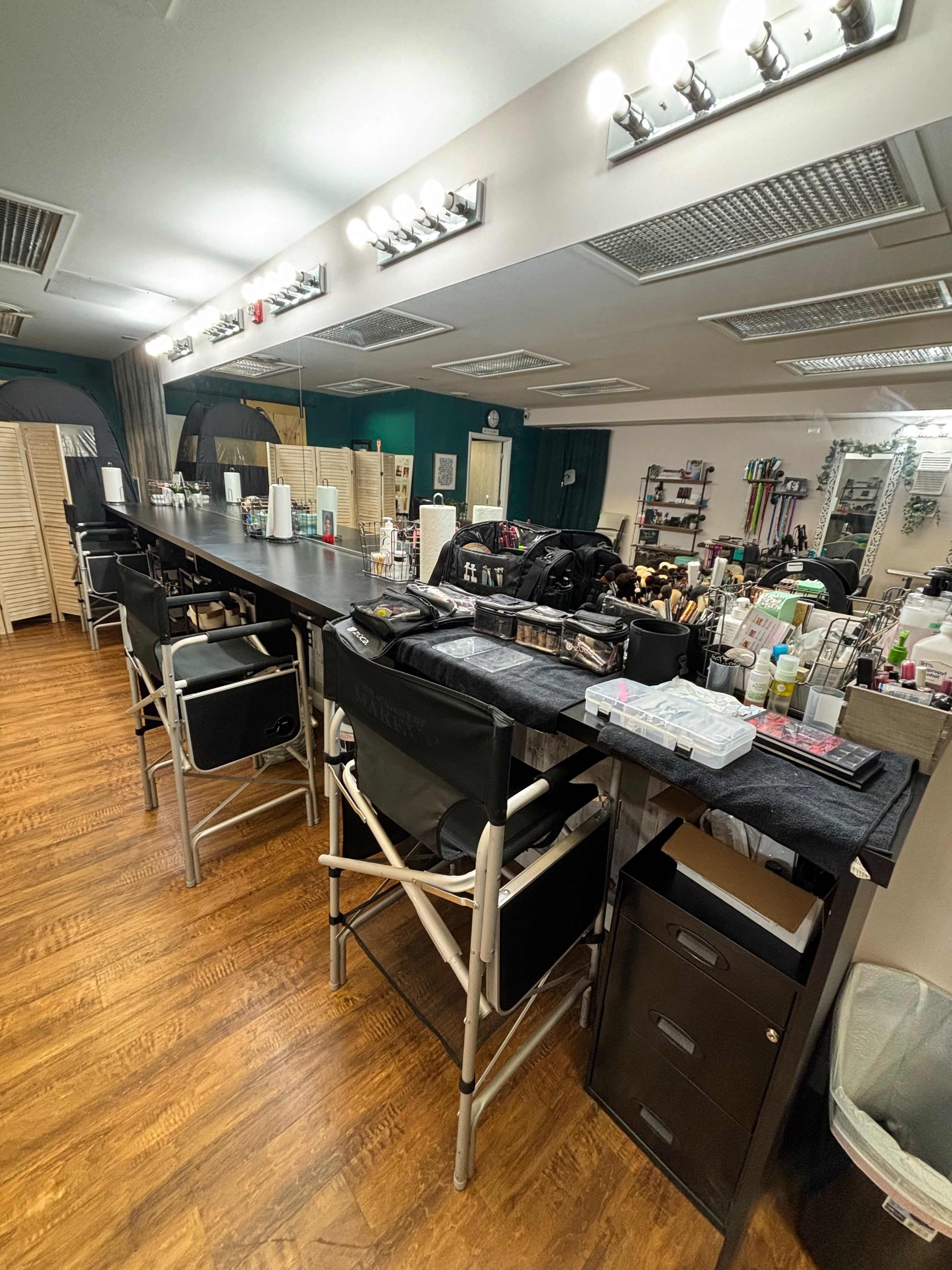 The image shows a spacious beauty salon with a long, dark countertop lined with makeup tools and supplies, surrounded by director's chairs on one side.