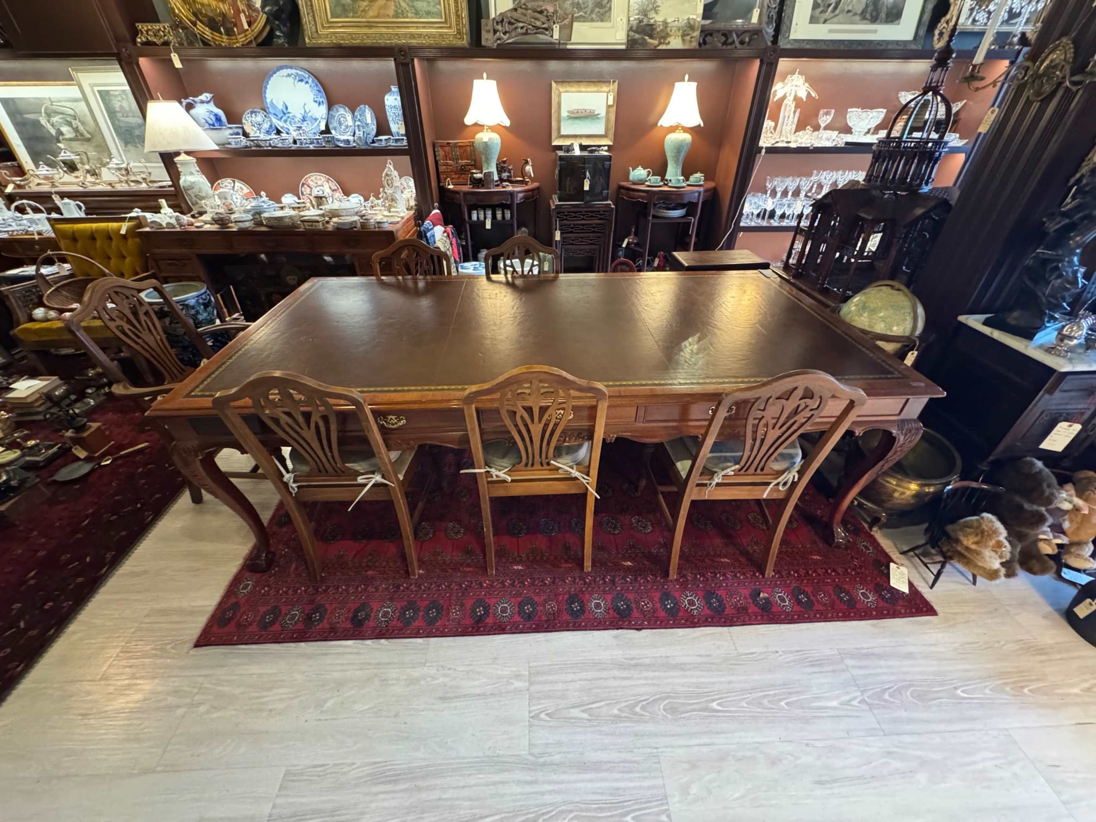 A large wooden dining table with a dark leather surface is surrounded by four matching chairs in a vintage shop filled with various antiques and collectibles.