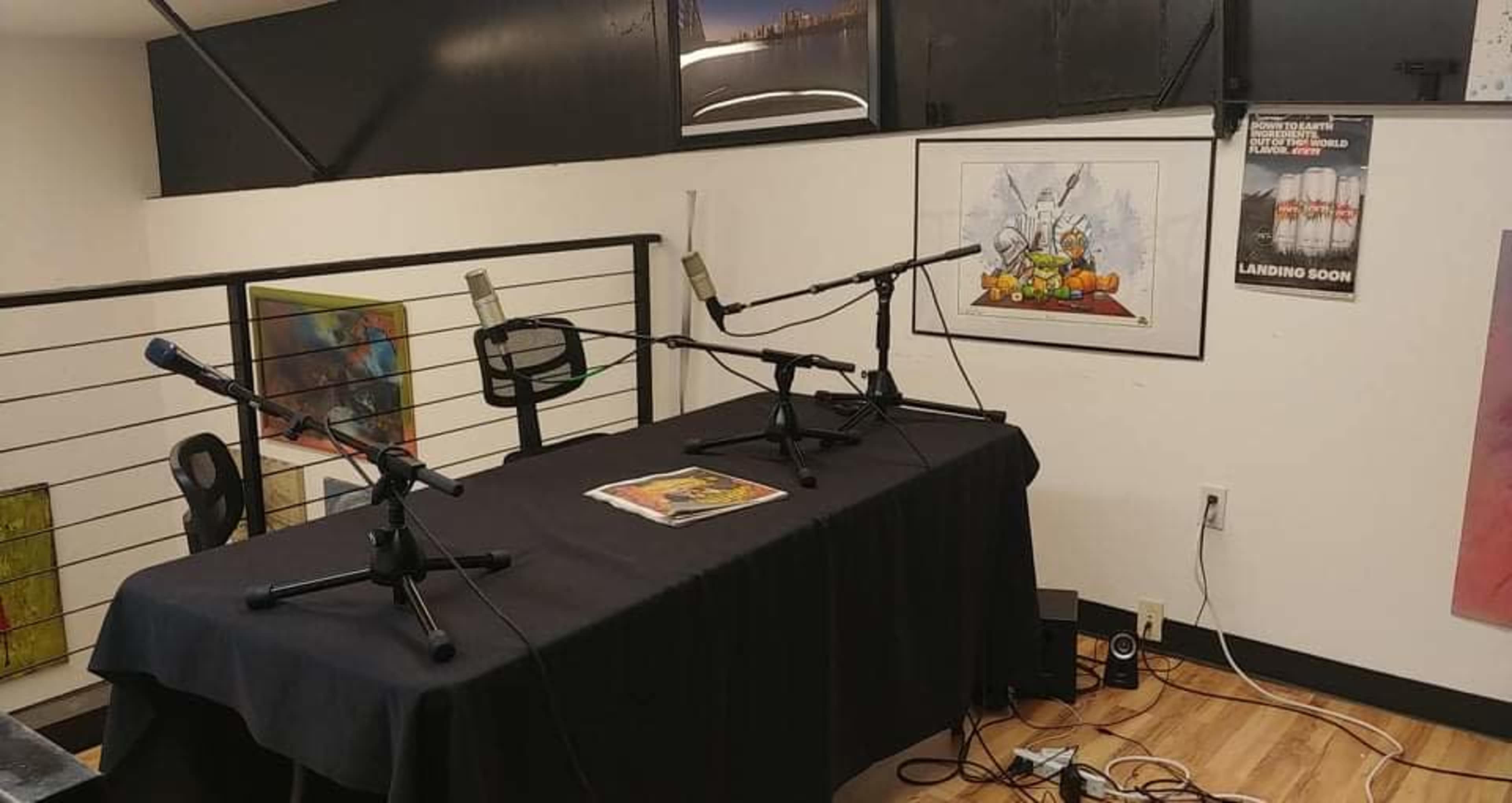 A table with a black tablecloth and three microphones is set up in a room with artwork on the walls and a speaker on the floor.
