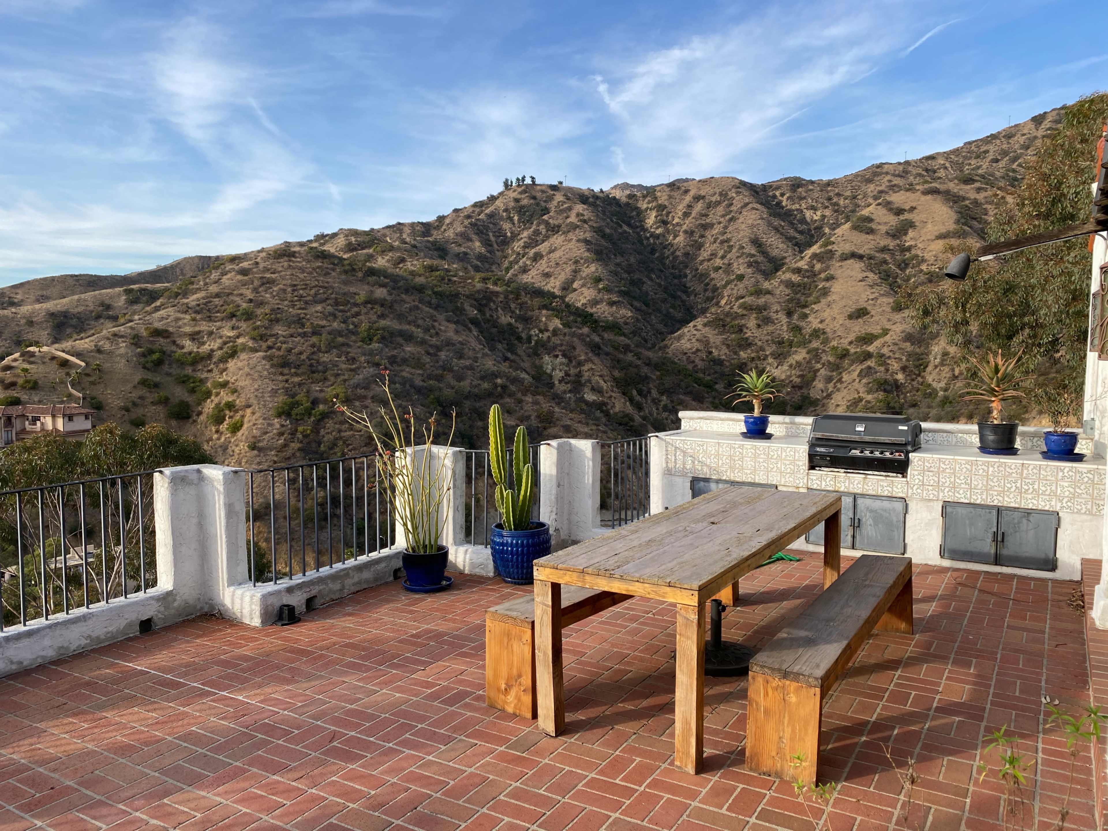 A wooden table and benches are set on a brick patio overlooking a hilly landscape with sparse vegetation.