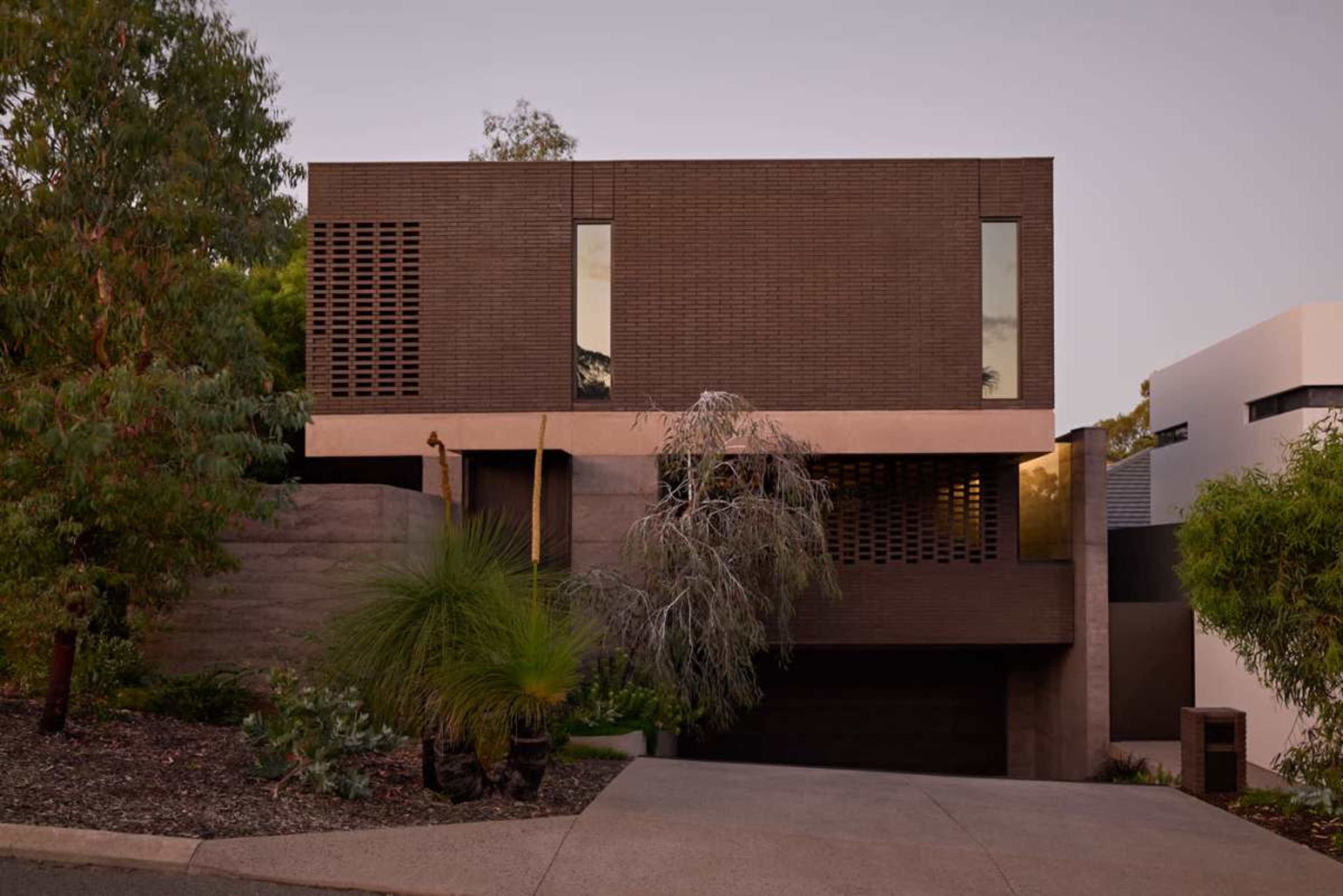 A modern two-story house features a combination of dark brick and concrete with large windows set in a landscaped setting.
