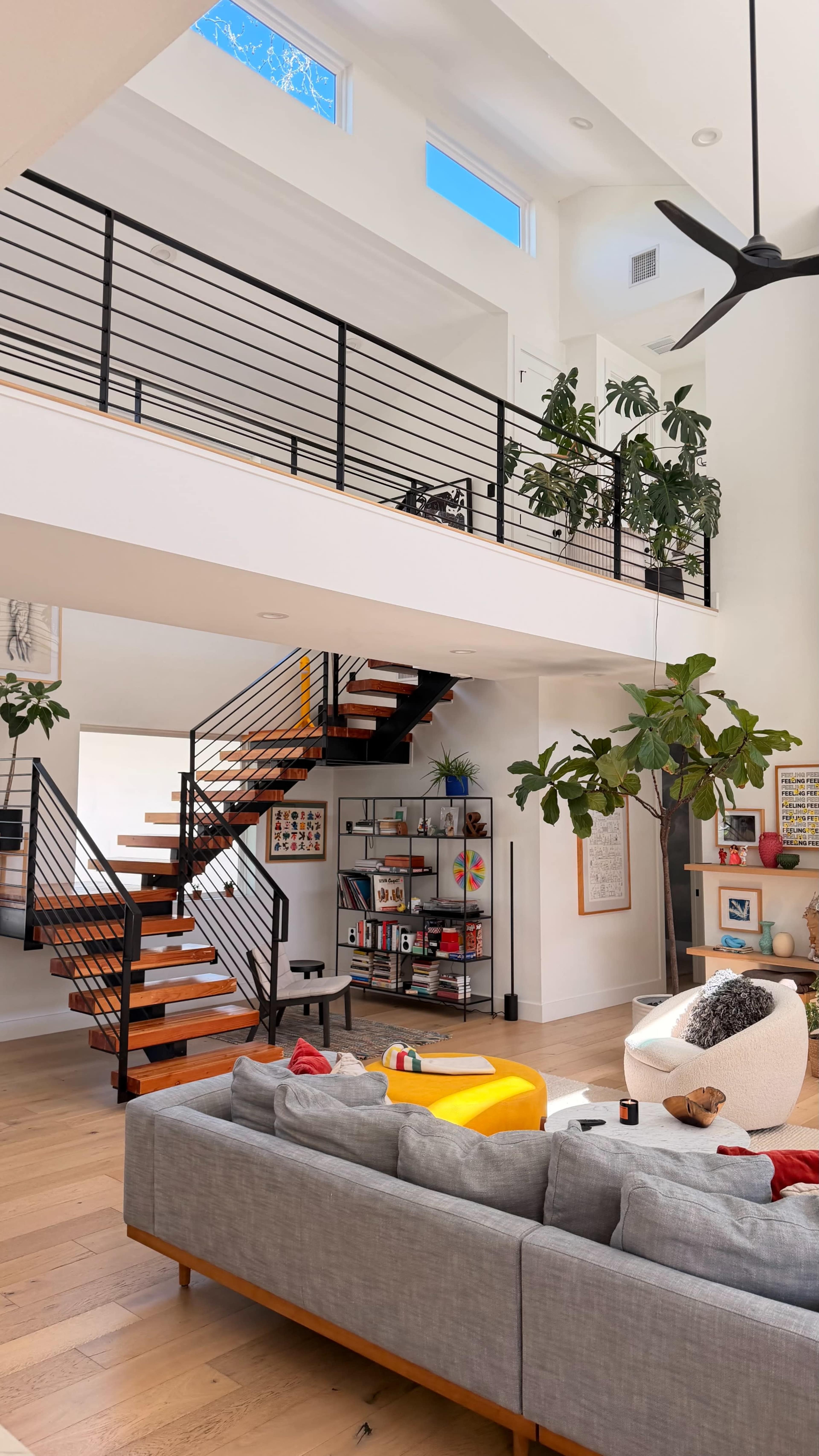 The image shows a modern interior design featuring an open staircase, a spacious living area with a gray sofa, and decorative plants throughout.
