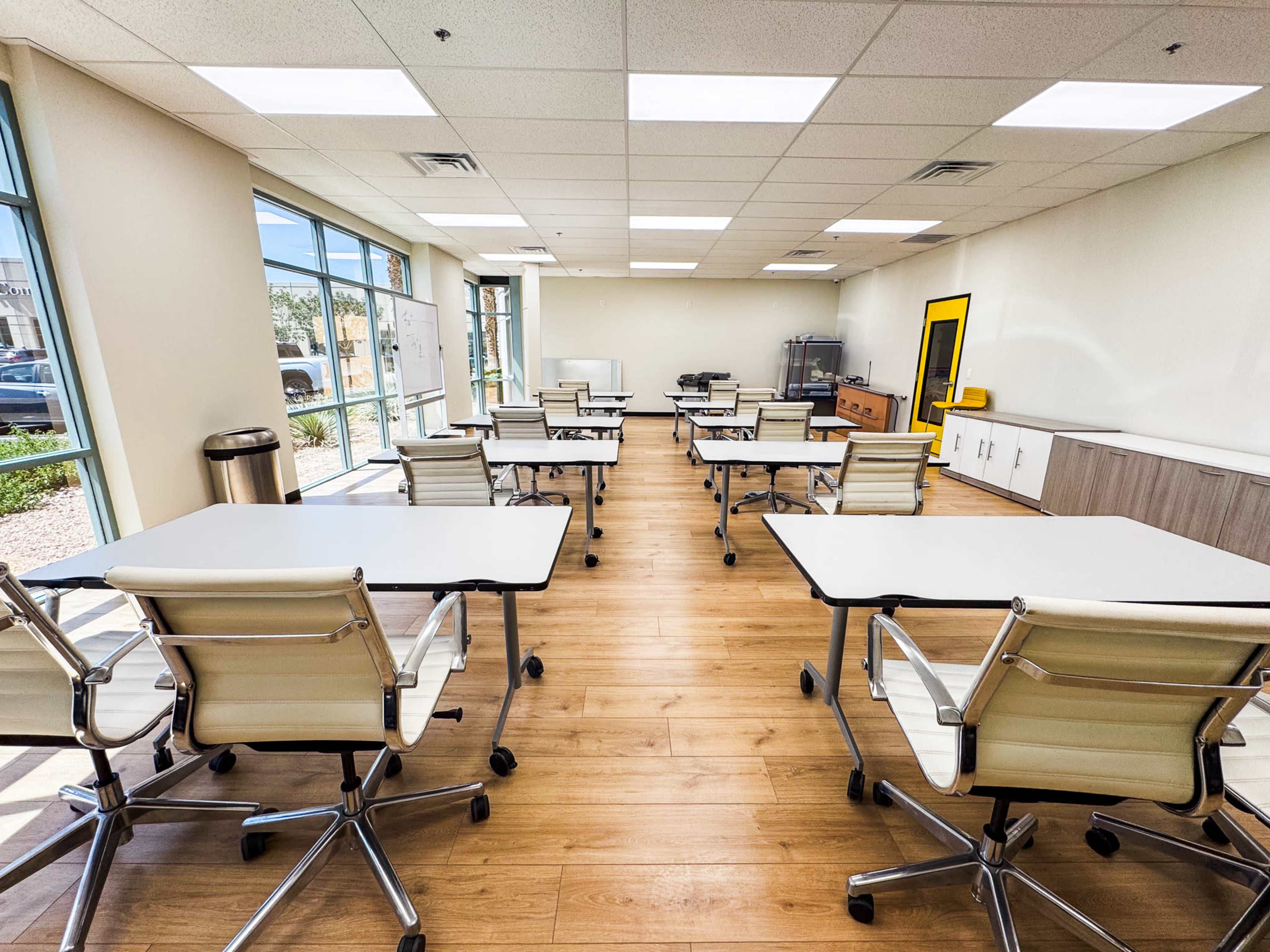The image shows a modern classroom with multiple tables and chairs arranged in rows, windows allowing natural light, and a combination of whiteboards and wooden cabinetry along the walls.