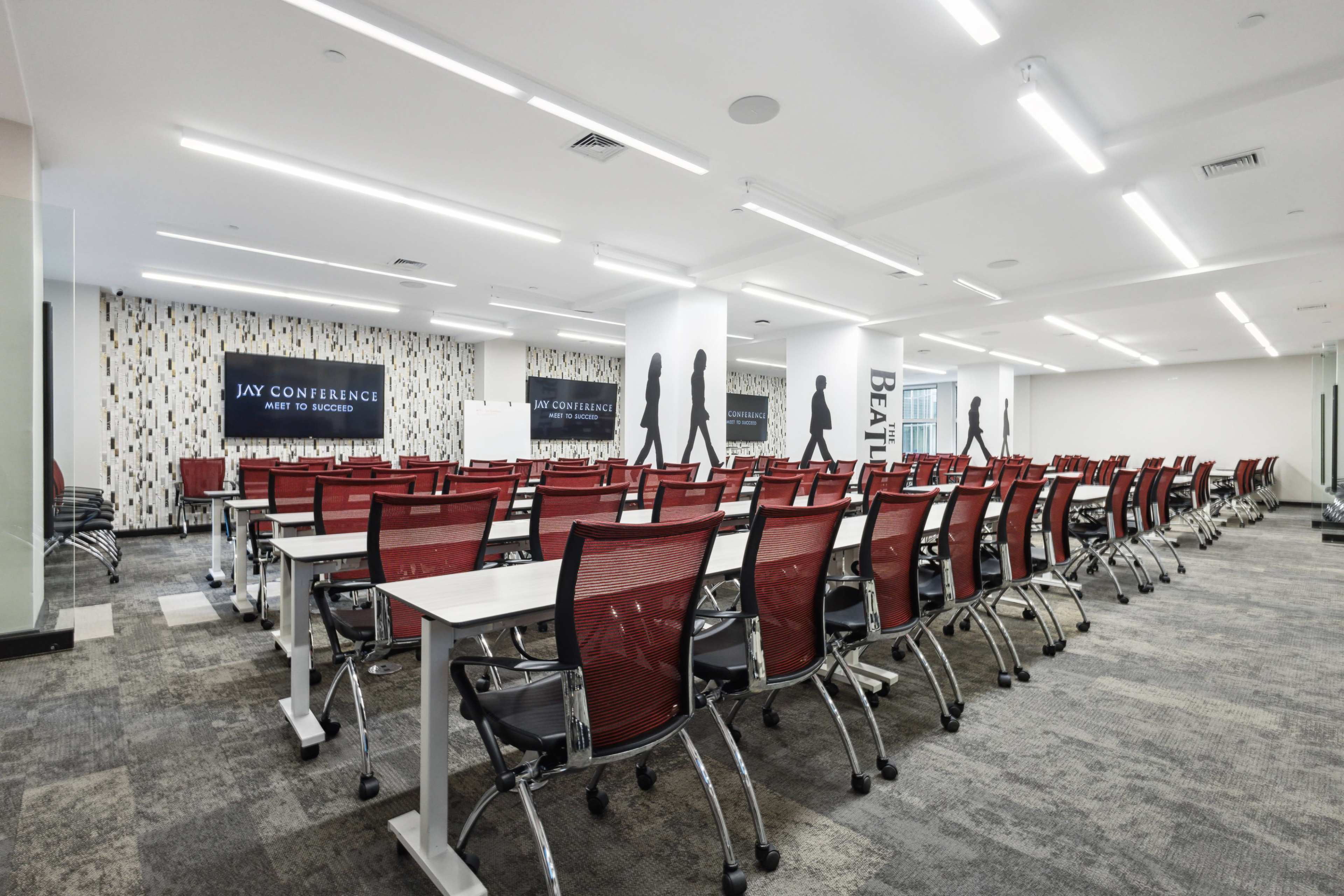 A modern conference room features rows of red seated tables arranged for a meeting, with wall graphics and ample overhead lighting.