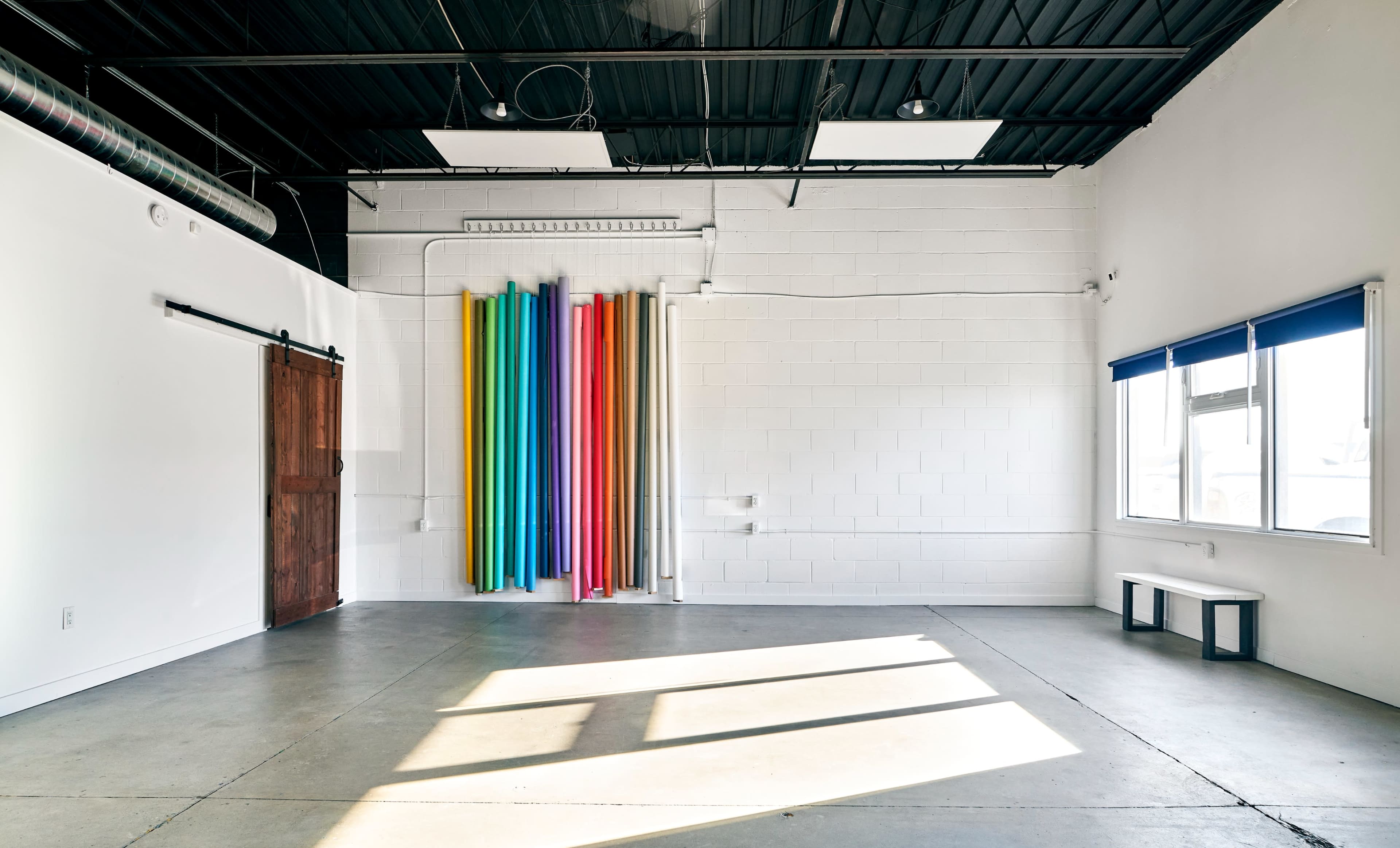 The image shows a spacious, empty room with a concrete floor, featuring colorful rolls of paper hanging on the wall and a wooden sliding door.