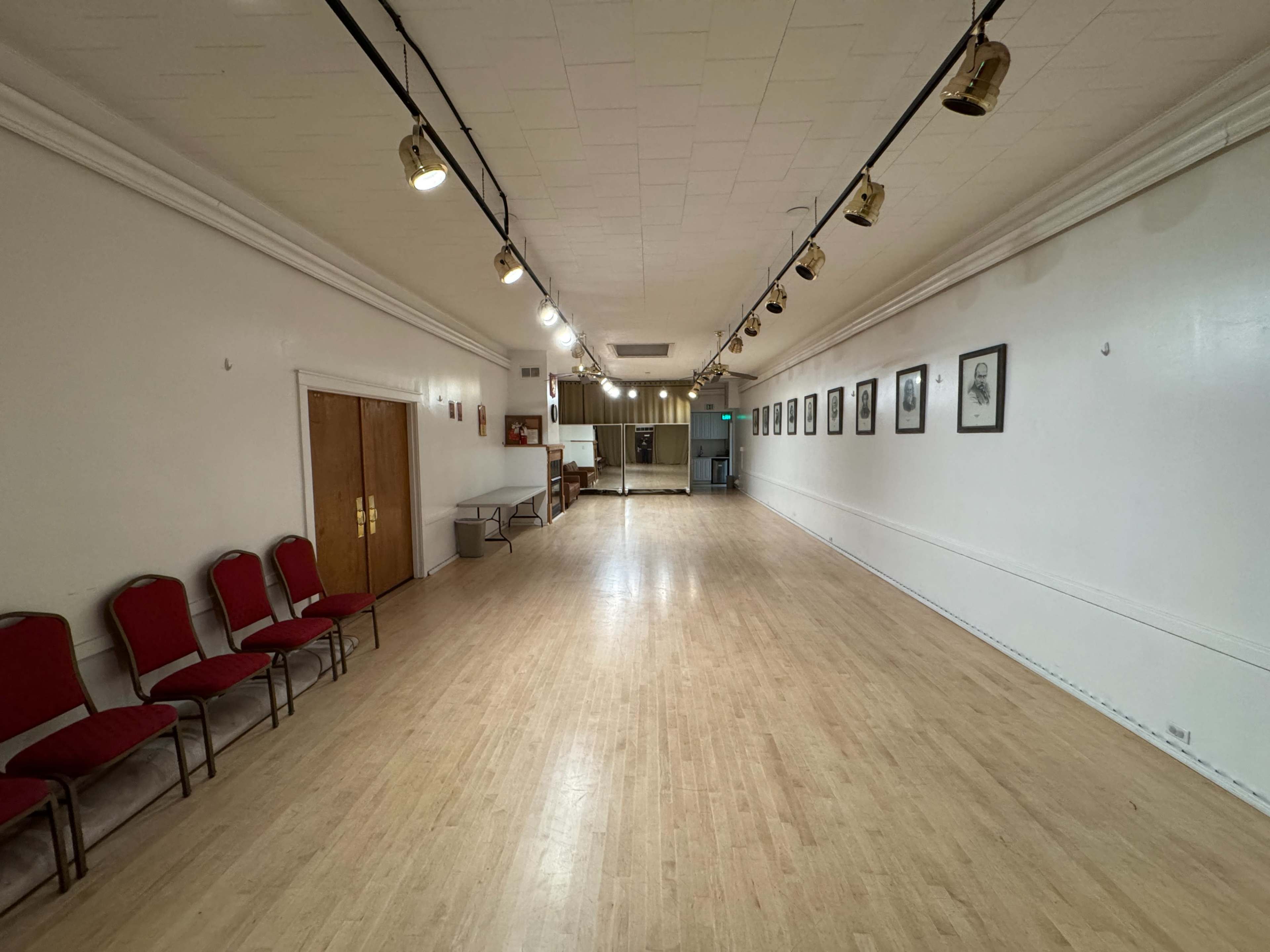 The image shows a spacious room with a wooden floor, lined with several red chairs on one side and framed pictures on the white walls.