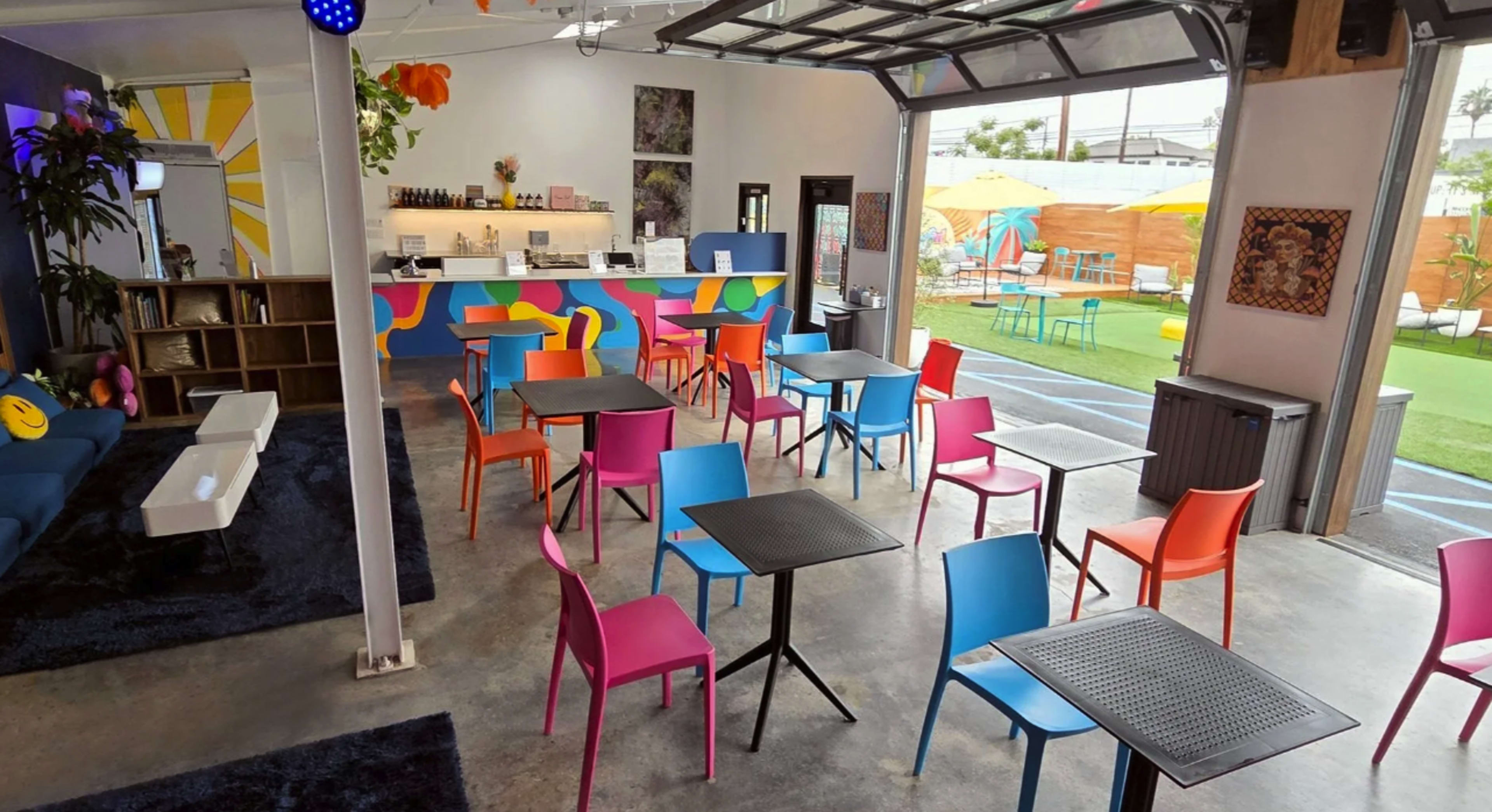 The image shows a modern space featuring colorful chairs and tables arranged around a bar area with a vibrant mural, and an outdoor seating area visible through large glass doors.