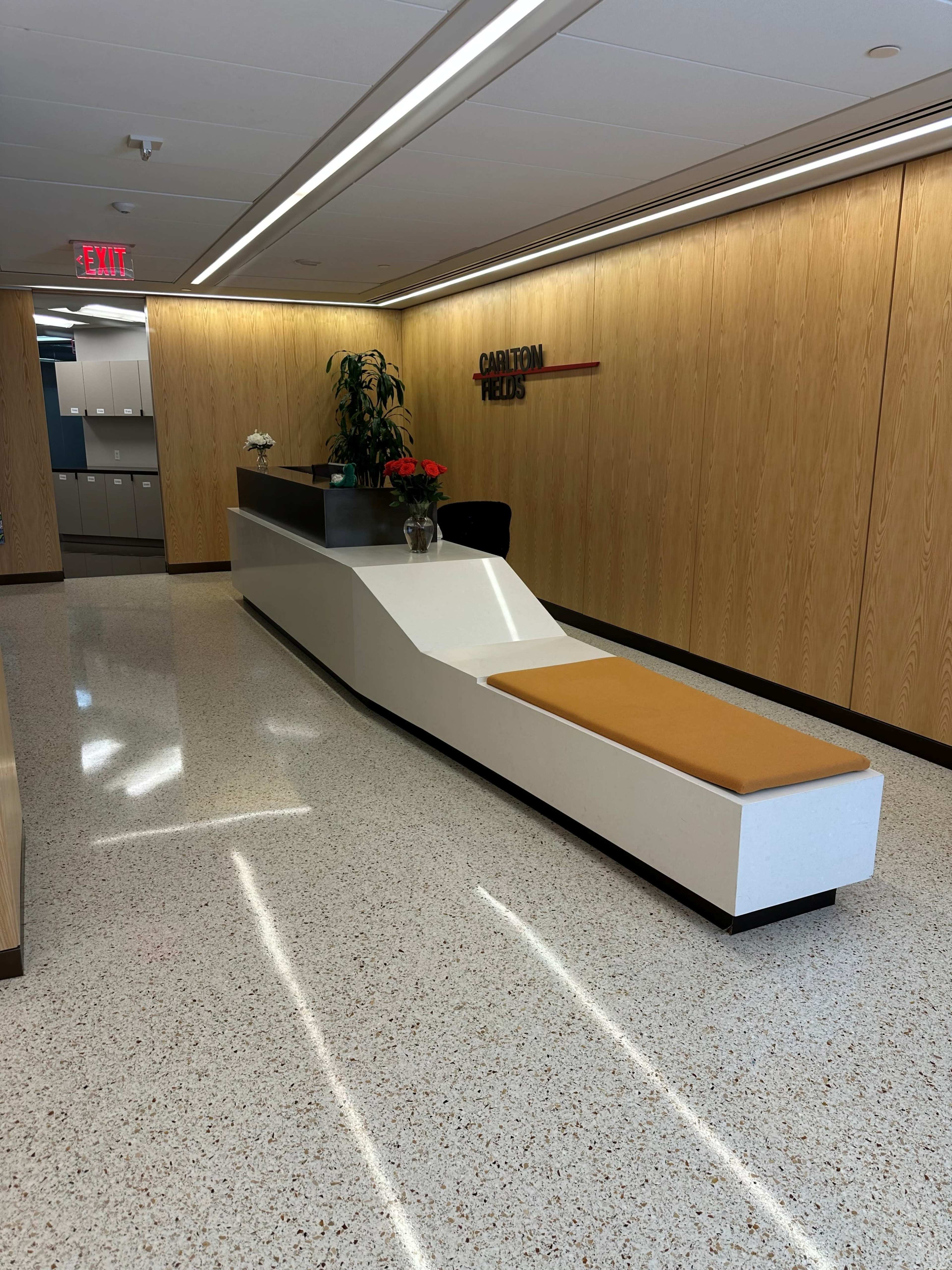 The image shows a modern office reception area with a sleek white reception desk and a long, cushioned seating area in front of it.