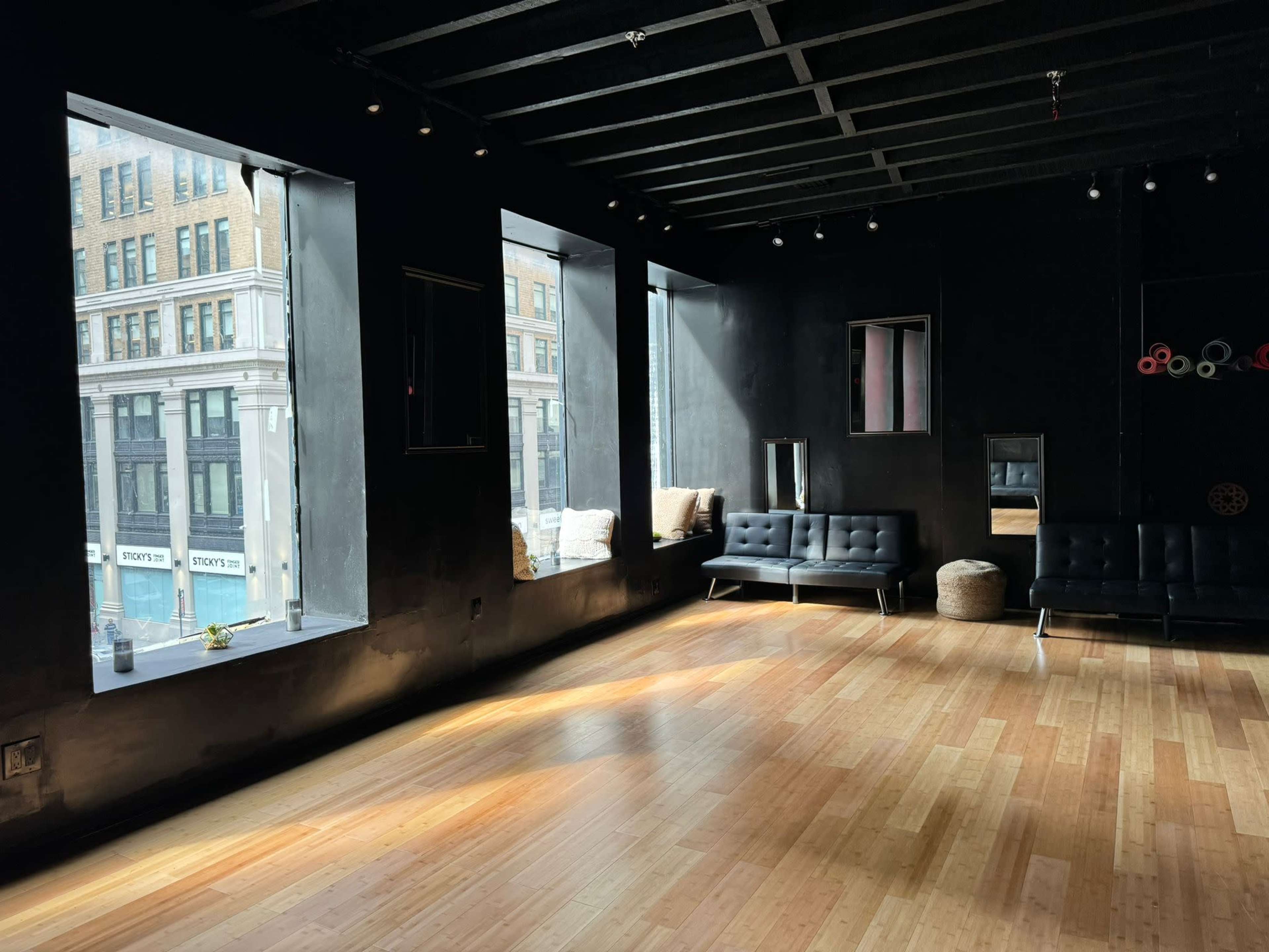 The room features large windows that allow natural light to illuminate a space with dark walls and wooden flooring, furnished with black couches and a few seating areas.