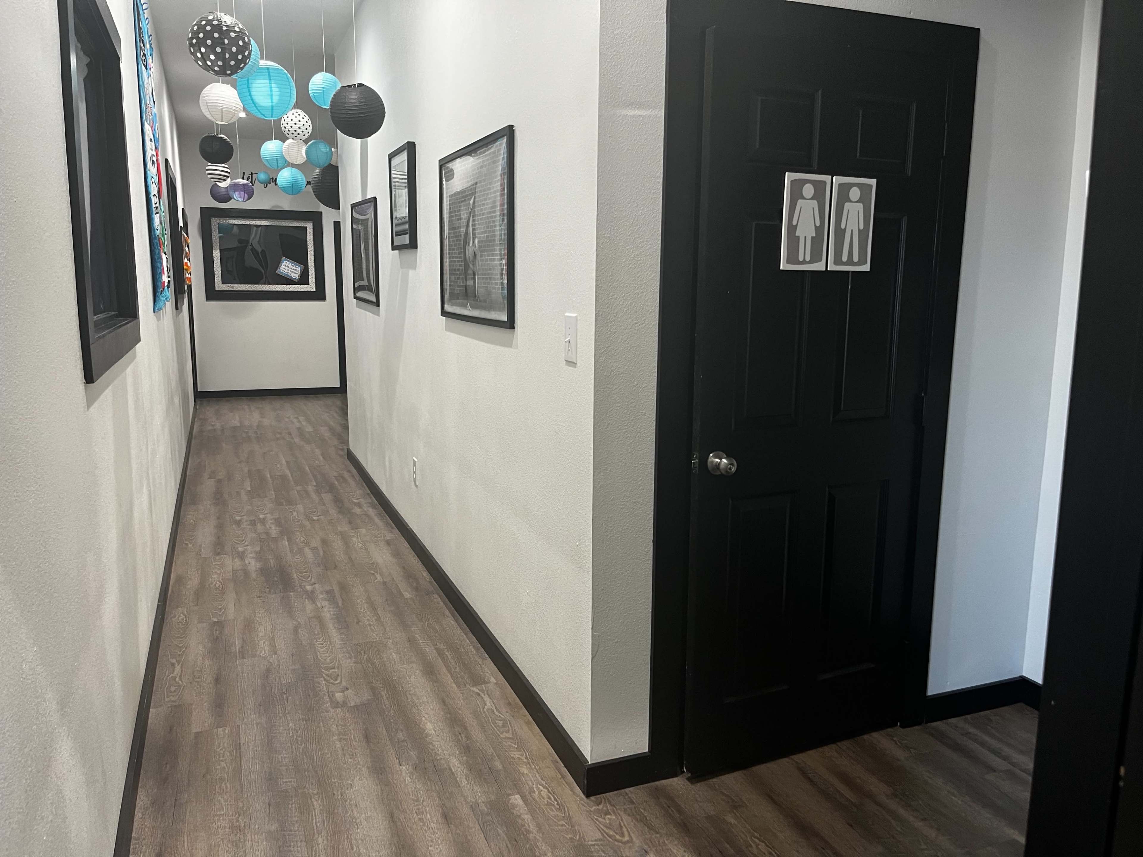 The image shows a long hallway with black doors leading to restrooms, framed pictures on the walls, and decorative hanging lamps.