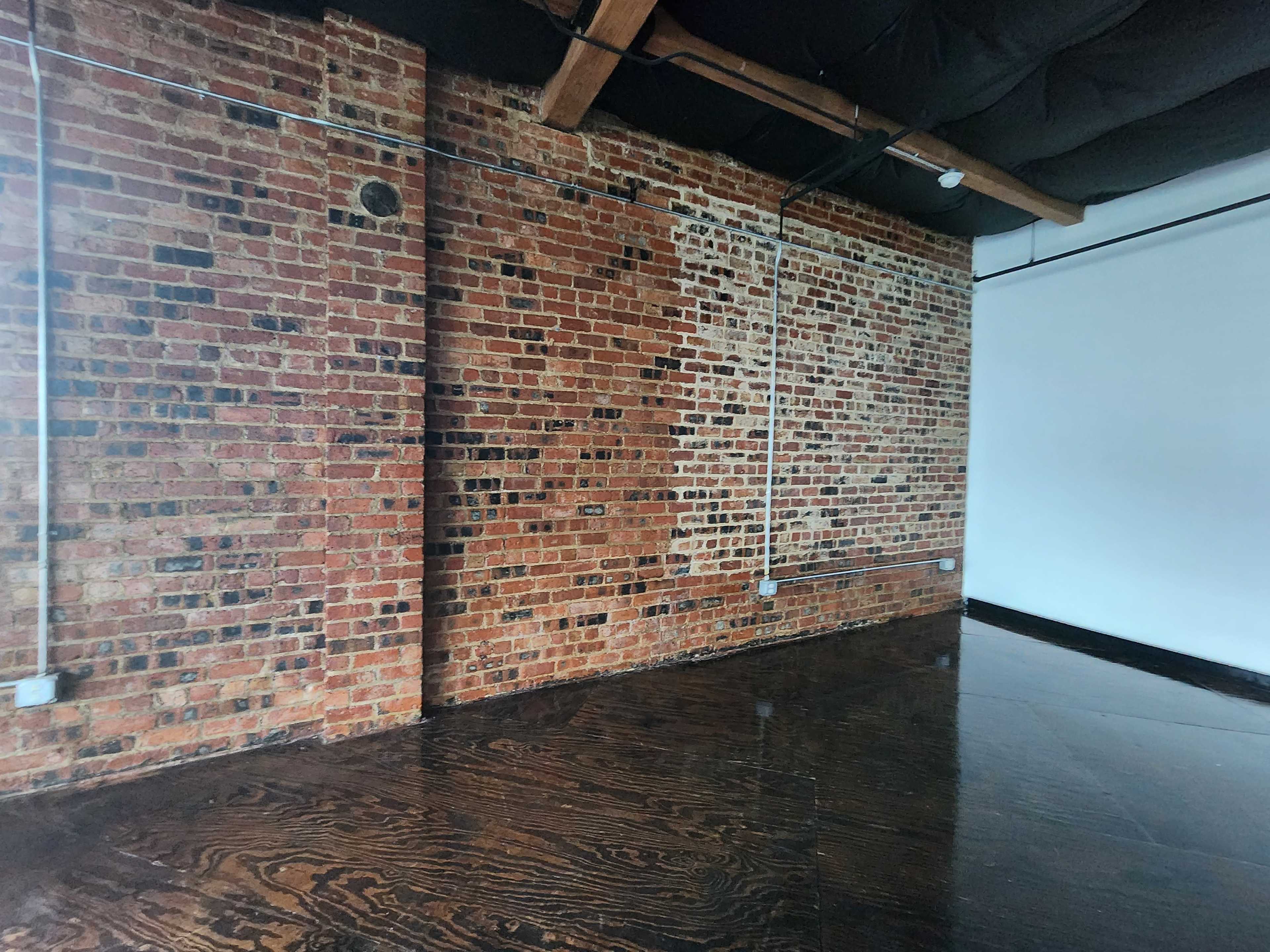 The image shows an interior space featuring a brick wall on one side and a white wall on the other, with dark wooden flooring.